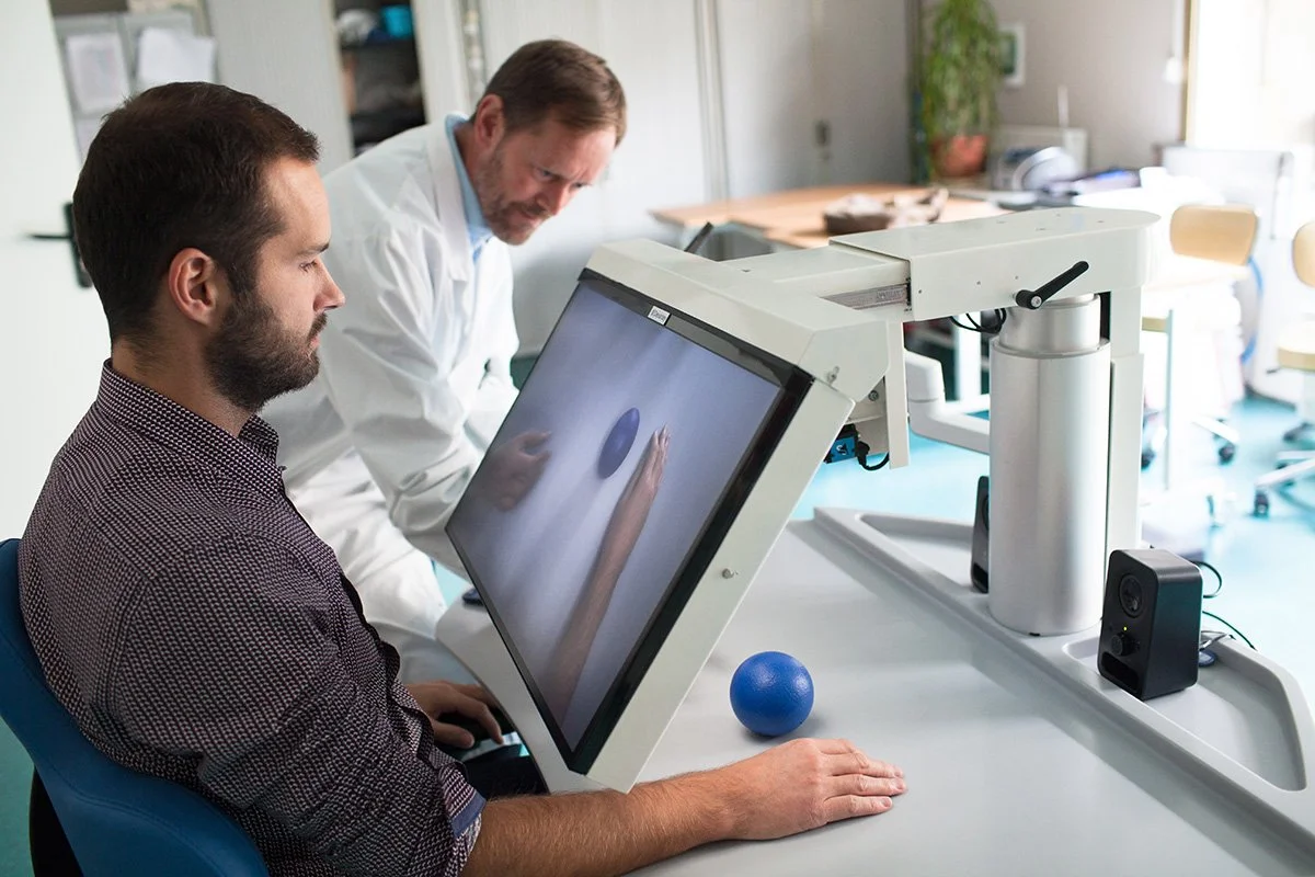 A man is sitting in front of a touchscreen interface with a medical or scientific device near him. A doctor or scientist is standing beside him, observing the screen. The environment is a clinical or laboratory setting with various equipment and furniture in the background.