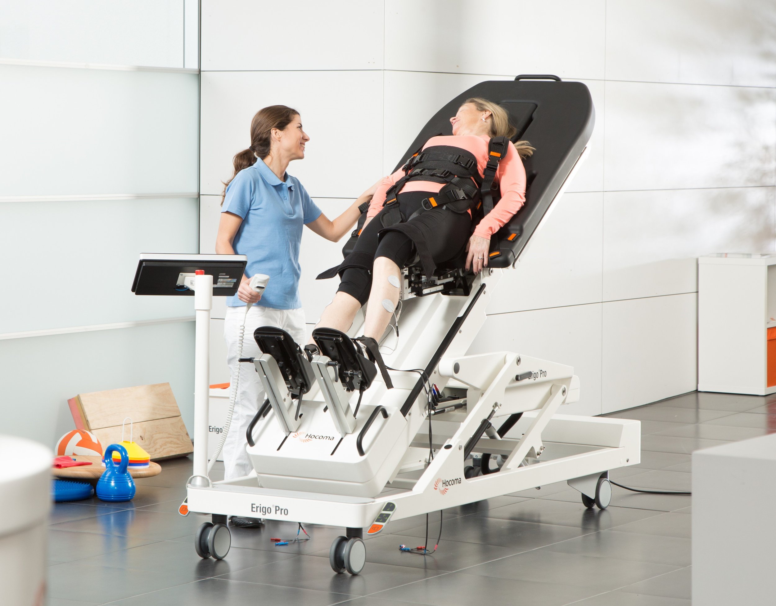 A hospital room with a woman lying on a medical stretcher, smiling and talking to a healthcare provider. The woman appears to be resting or recovering, with medical patches on her leg. The healthcare provider is holding a small device and wearing a blue uniform. The room has a light-colored wall, some toys on a wooden board, and medical equipment nearby.