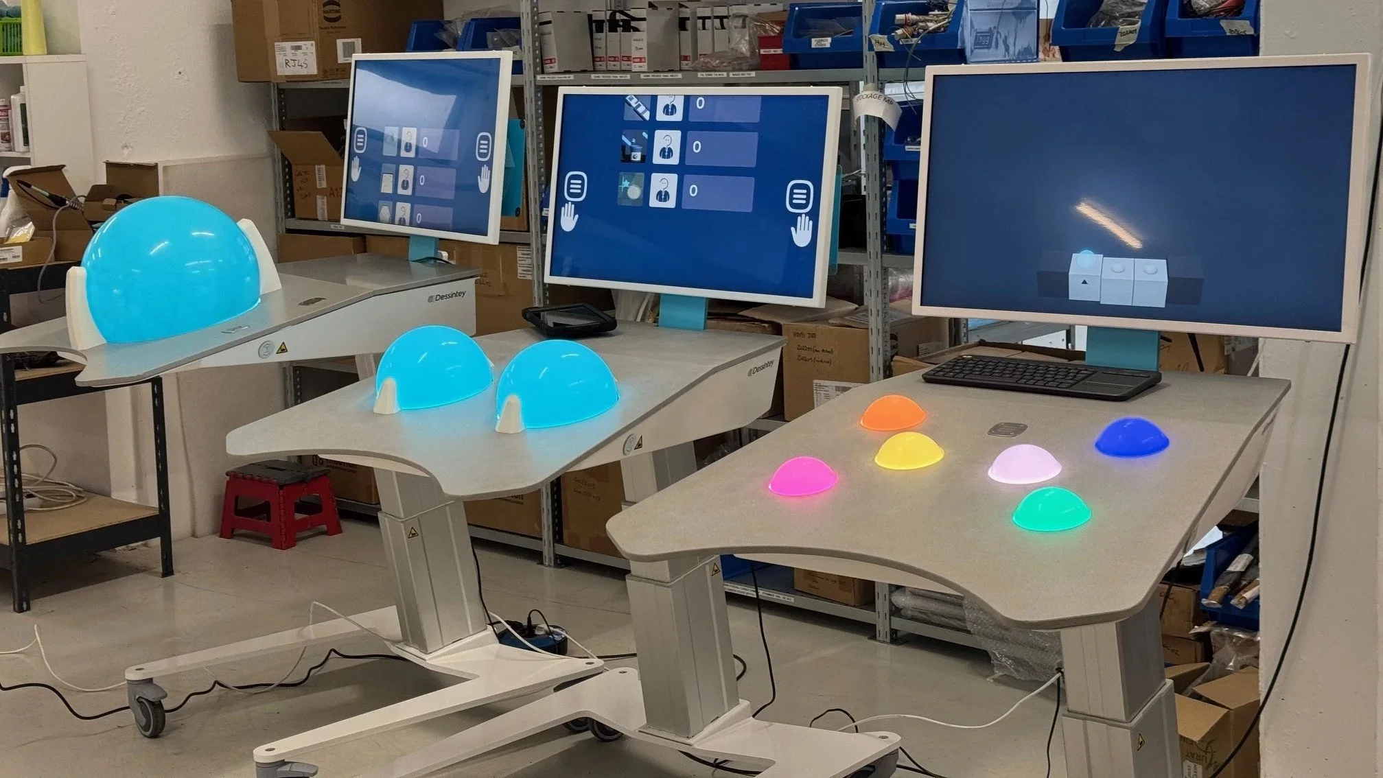Photo of a workspace with three computer monitors, some colorful illuminated dome-shaped objects on the tables, and scientific equipment including a blue hemisphere with a support structure, with shelves and boxes in the background.