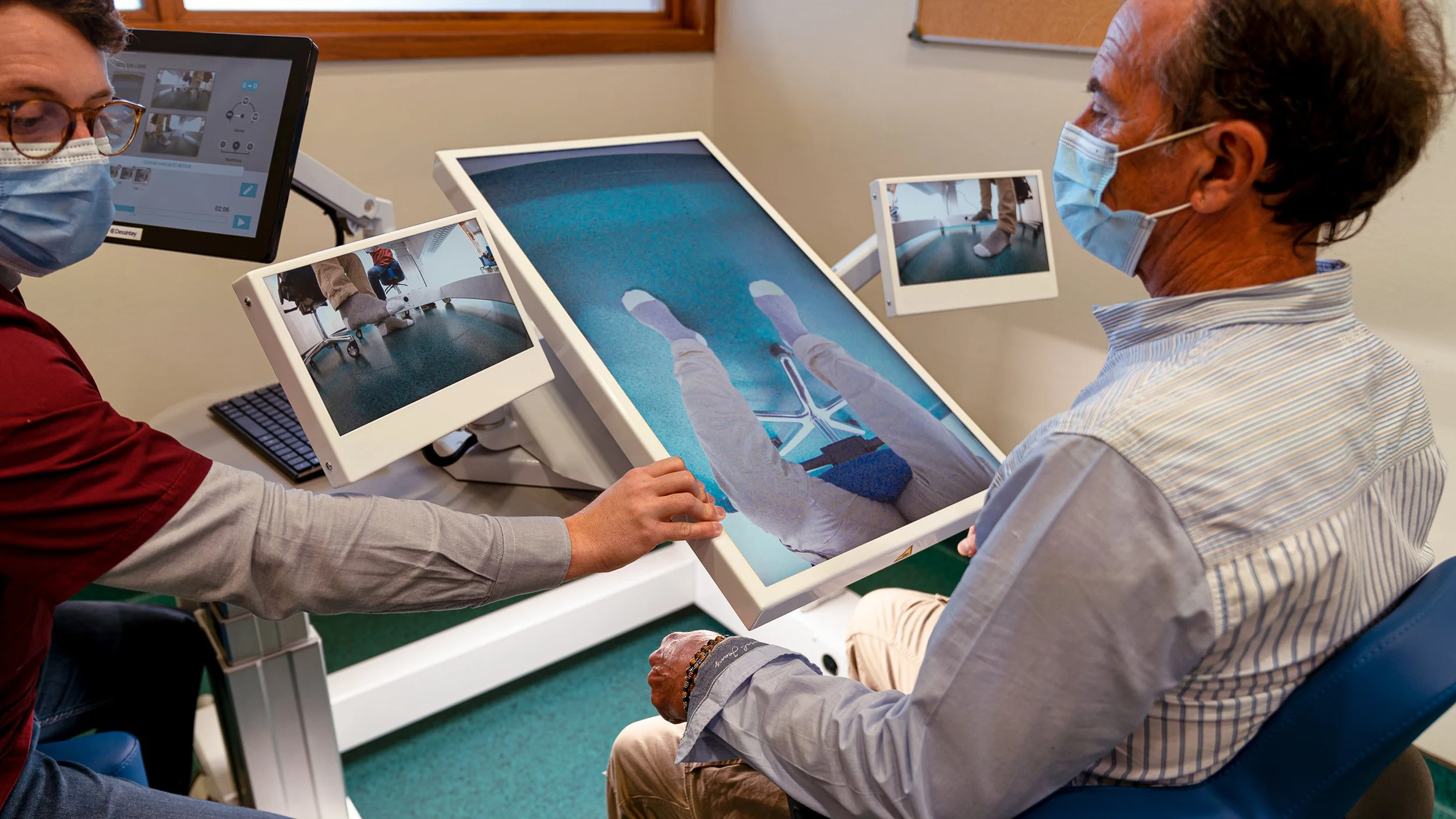 A healthcare professional conducting a virtual reality therapy session with an elderly man wearing a mask. The setup includes multiple screens showing the man's feet and legs using virtual reality technology.