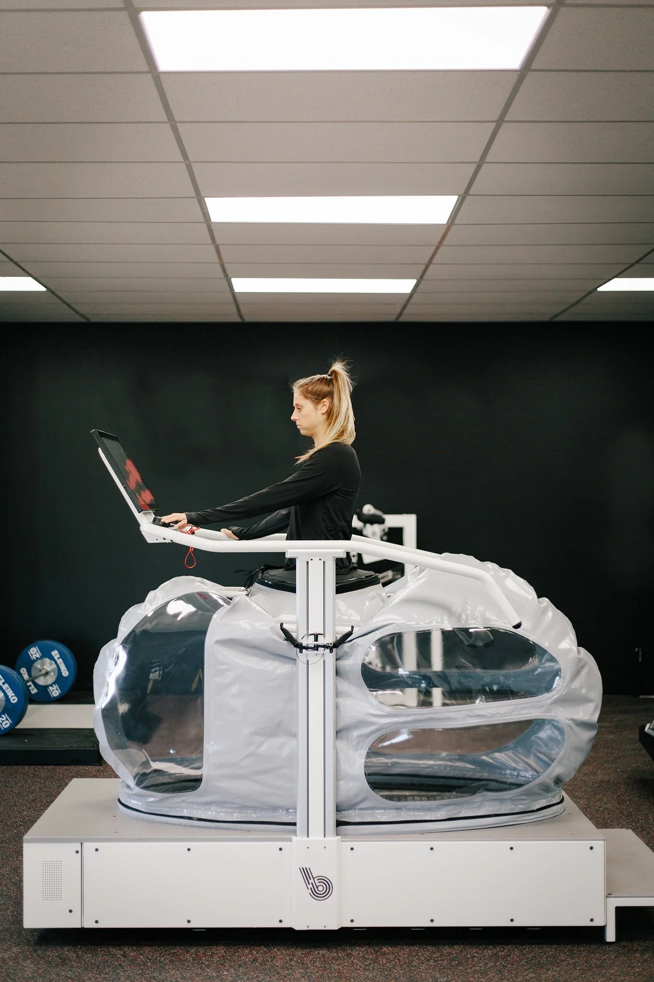 A woman working on a virtual reality simulator designed to look like a futuristic car, with a clear plastic exterior and sleek design, in a room with black walls and ceiling lights.
