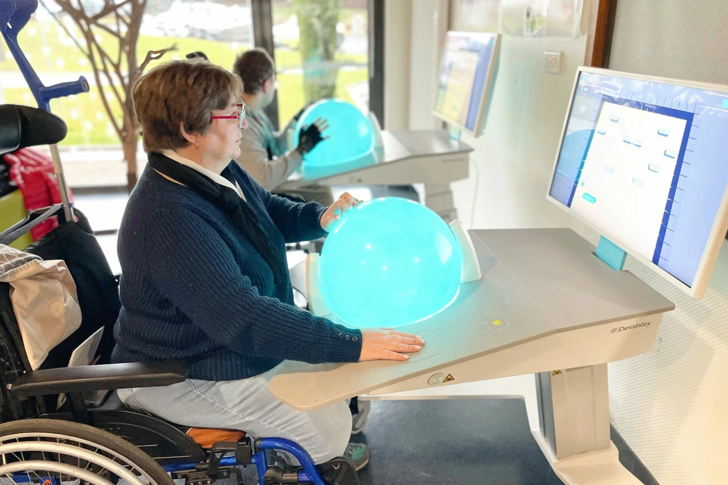 A woman in a wheelchair is sitting at a desk with a glowing blue globe in front of her, and she is touching the globe. In the background, a young girl is sitting at a desk with her hand on a blue globe, and there are computer monitors on the desks. The environment appears to be a classroom or learning center with large windows.