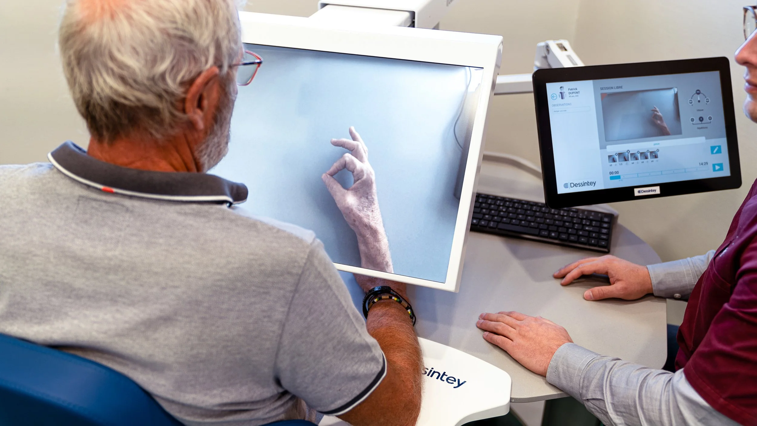 An elderly man with white hair and glasses sitting at a table, using a touch screen device for a medical or psychological assessment, with a professional woman assisting and a computer monitor displaying the session interface.