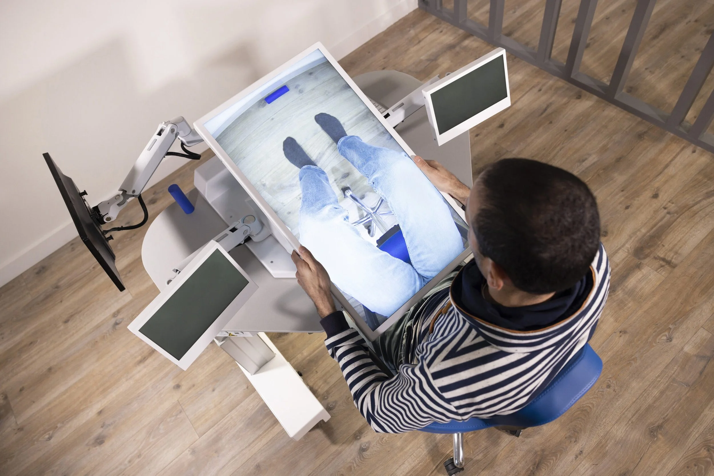 A person sits in a blue chair using a tilt table equipment to stand upright. The person's legs are visible on a screen, showing the view from their perspective.