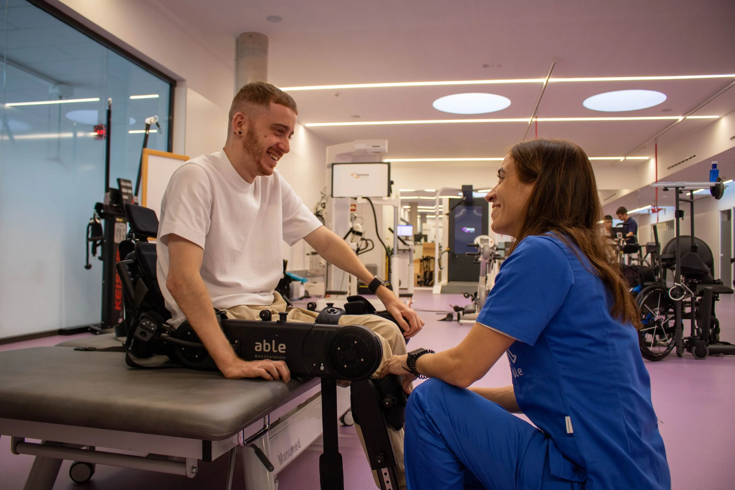 A young man in a wheelchair smiling and holding hands with a woman who is kneeling, in a modern healthcare or rehabilitation center, engaging in a therapy session.