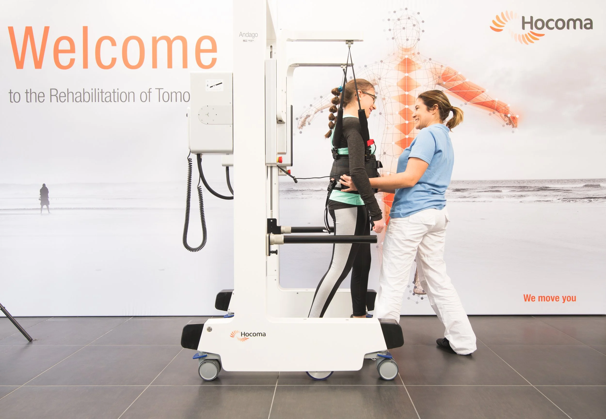 Two women in a medical rehabilitation facility are using a standing frame to support a patient during therapy. One woman, wearing glasses and sportswear, is in the standing frame, smiling and engaging with another woman in scrubs. The background displays an anatomical diagram with the words 'Welcome' and 'Hocoma,' indicating a facility focused on patient rehabilitation.