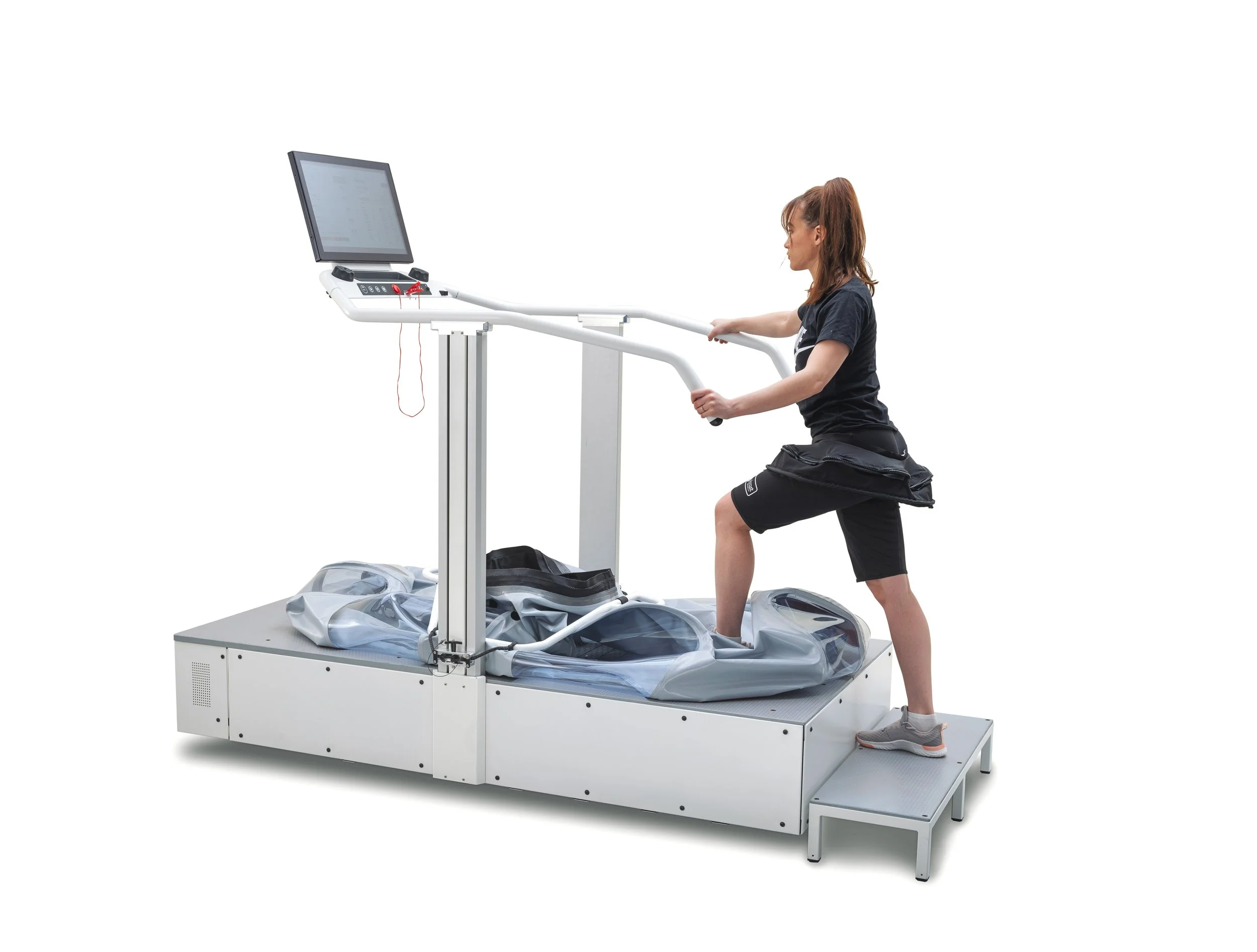 A woman in black workout clothing is kneeling on one knee while using a medical exercise device with a monitor, white frame, and a platform, isolated on a plain white background.