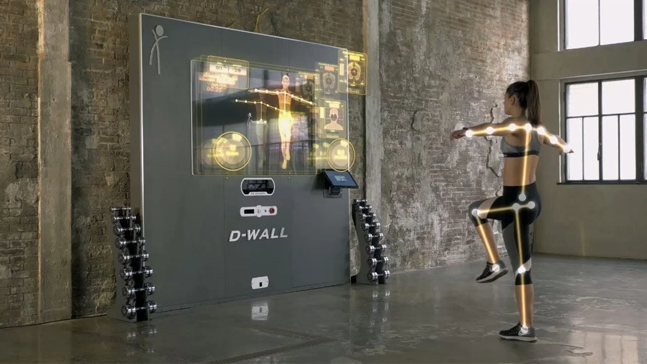 A woman in workout clothes is performing a balance exercise in a fitness lab with high-tech fitness equipment. The lab has exposed brick walls, large windows, and an interactive screen displaying biofeedback data.