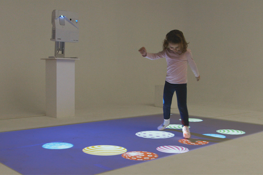 A girl in a pink shirt and dark pants jumps on an interactive floor with colorful, illuminated circle patterns in a neutral room. A projector on a stand is projecting the patterns onto the floor.