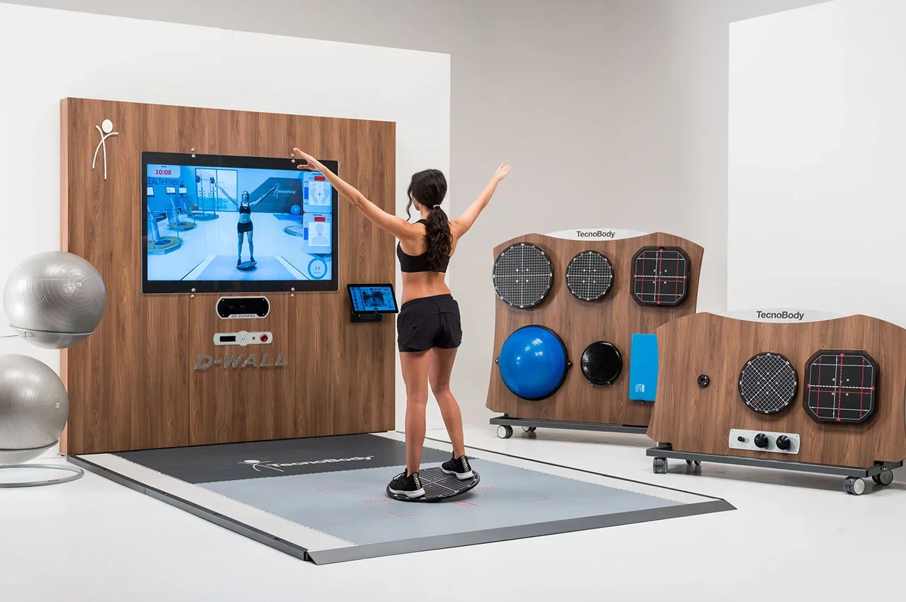 Woman in athletic attire using a virtual reality exercise system with a large screen, exercise mats, and fitness equipment in a gym setting.