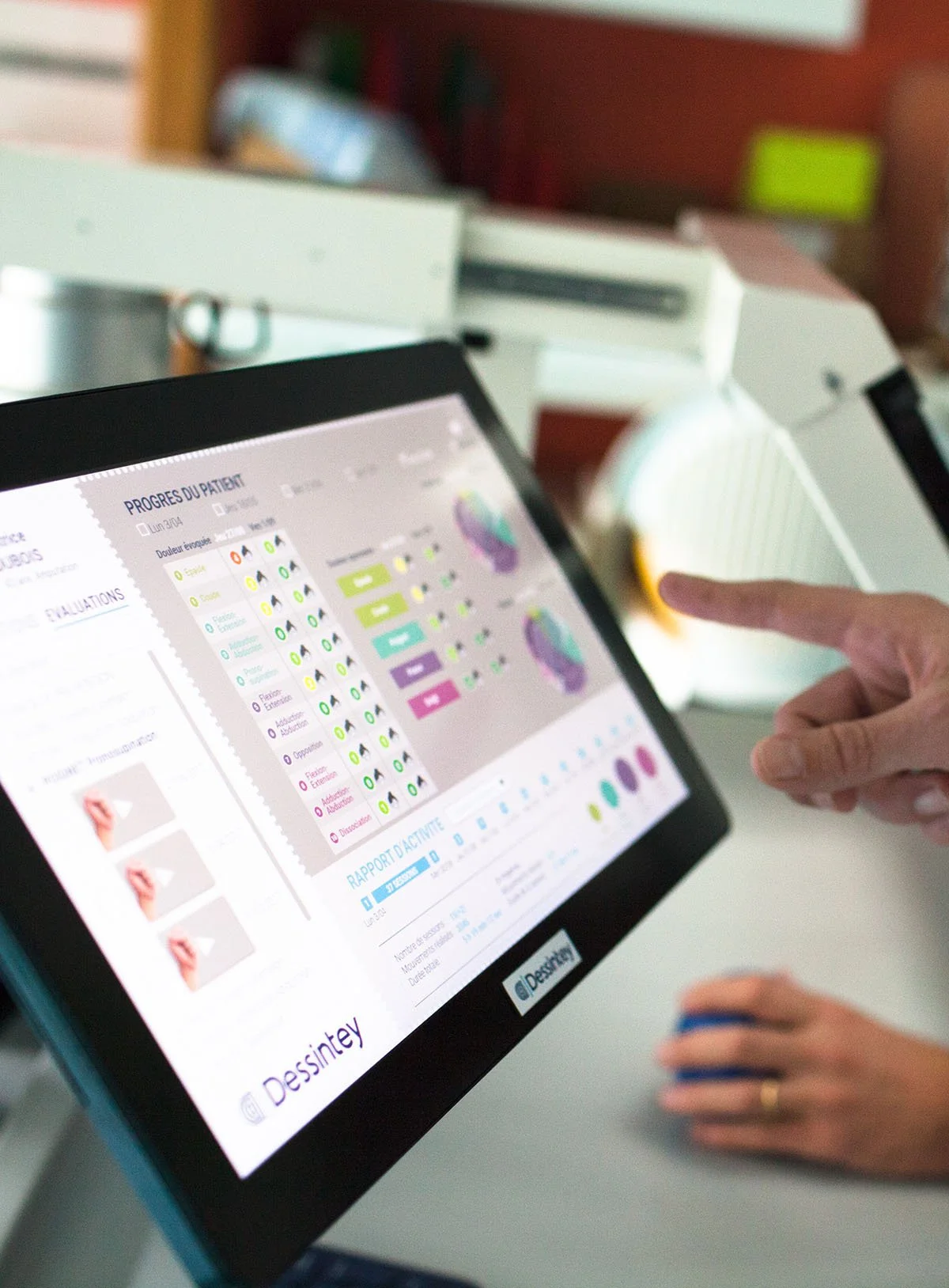 A person uses a touchscreen device to view a digital health or medical report, with a hand pointing at the screen. In the background, there is medical equipment, possibly a piece of diagnostic machinery.