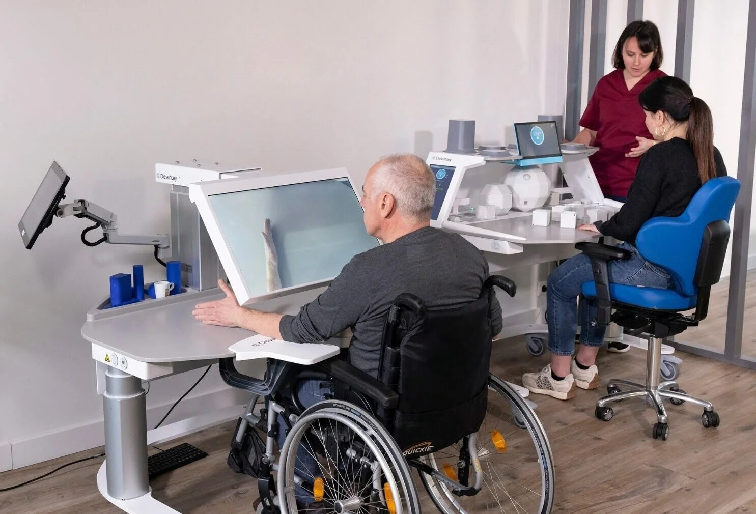 Two women and a man with a disability in a wheelchair in a healthcare or laboratory setting with medical and diagnostic equipment, including computers and monitors.