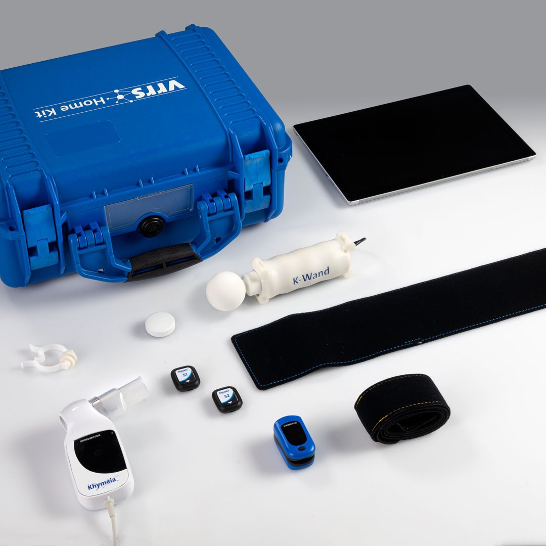 Medical equipment and supplies arranged on a white surface, including a blue hard case, a dark tablet, a white neural stimulation device labeled 'K-Wand', a rolled black strap, two small black devices with screens, a blue and black device, a white spray bottle, a black strap, and a white oval object.