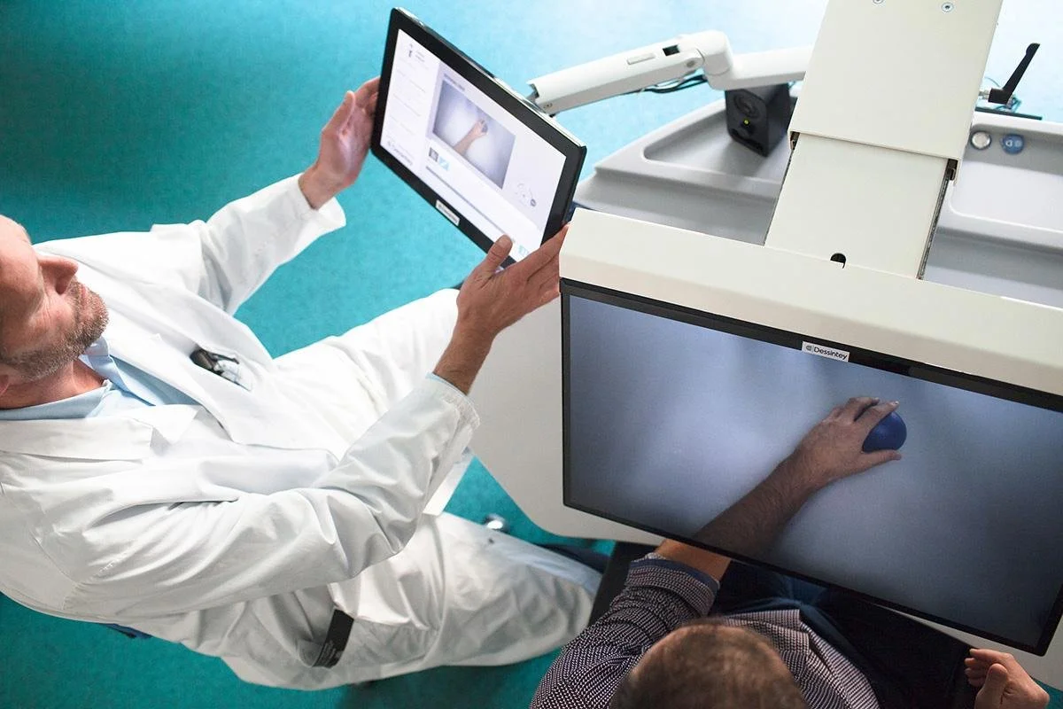 A doctor in a white coat sitting on the floor and holding a tablet, next to a medical scanner with a monitor and robotic arm, while another person uses a computer on a desk nearby.