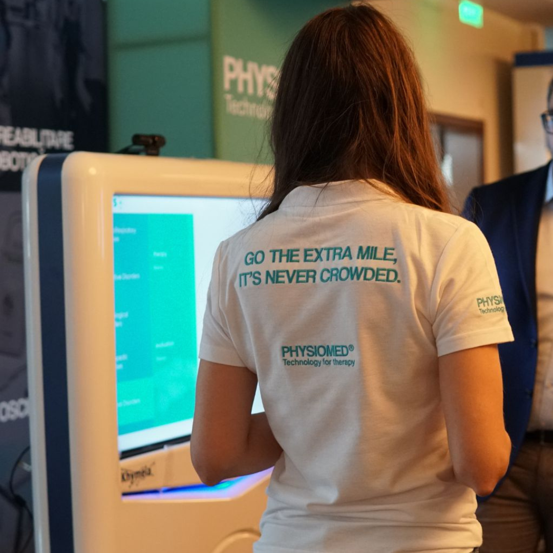 A woman with long brown hair wearing a white T-shirt with the text 'GO THE EXTRA MILE, IT'S NEVER CROWDED' on the back stands in front of a digital display at a conference or trade show. The booth or background features the brand 'PHYSIOMED'.