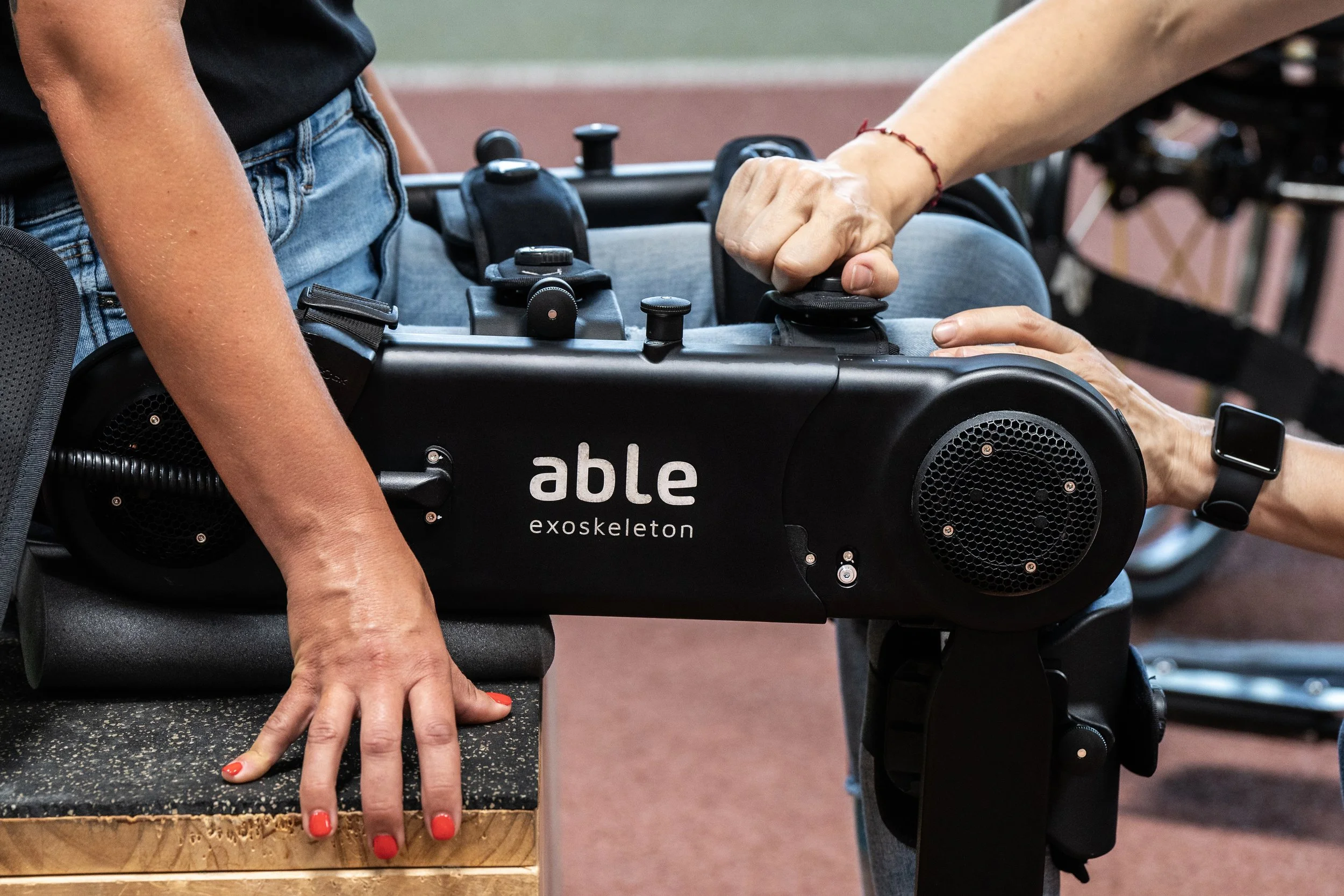 A person using an exoskeleton named Able to assist walking, with another person helping hold the device.
