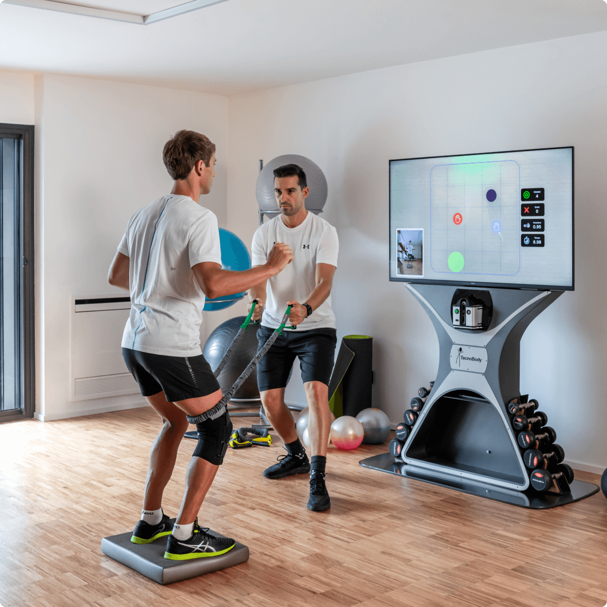 A man participating in a physical therapy session with a therapist in a modern therapy room, using a resistance band while standing on a balance pad. The room features exercise equipment, a large screen displaying a game or diagnostic interface, and a small video call window showing the man exercising.