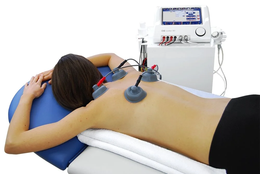 Person receiving electrotherapy treatment with multiple electrodes on their back, lying face down on a treatment table with a medical device in the background.
