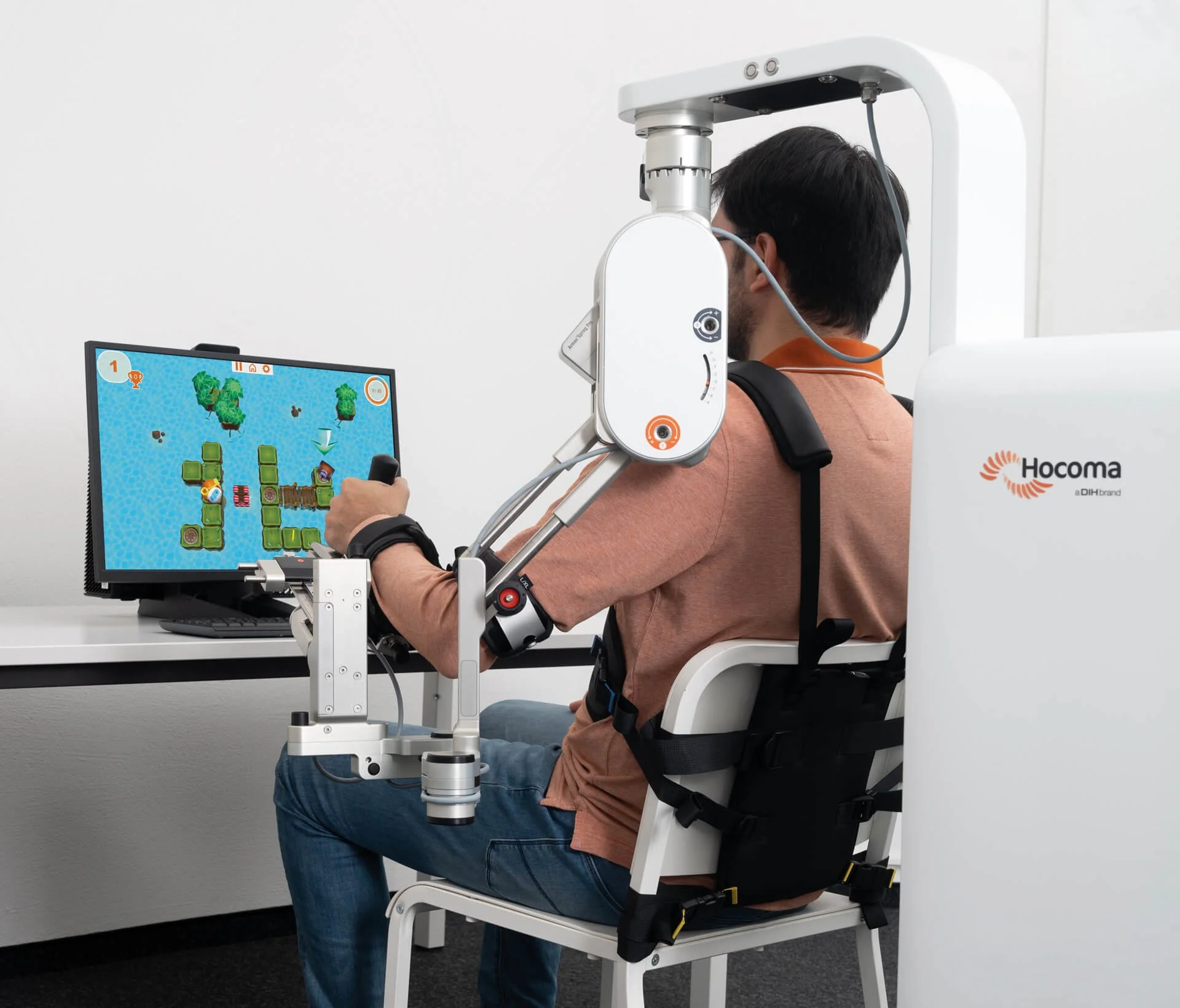 Person in a lab sitting in a specialized chair with a robotic device attached to their head, connected to a monitor displaying a colorful game or task involving trees and other objects.