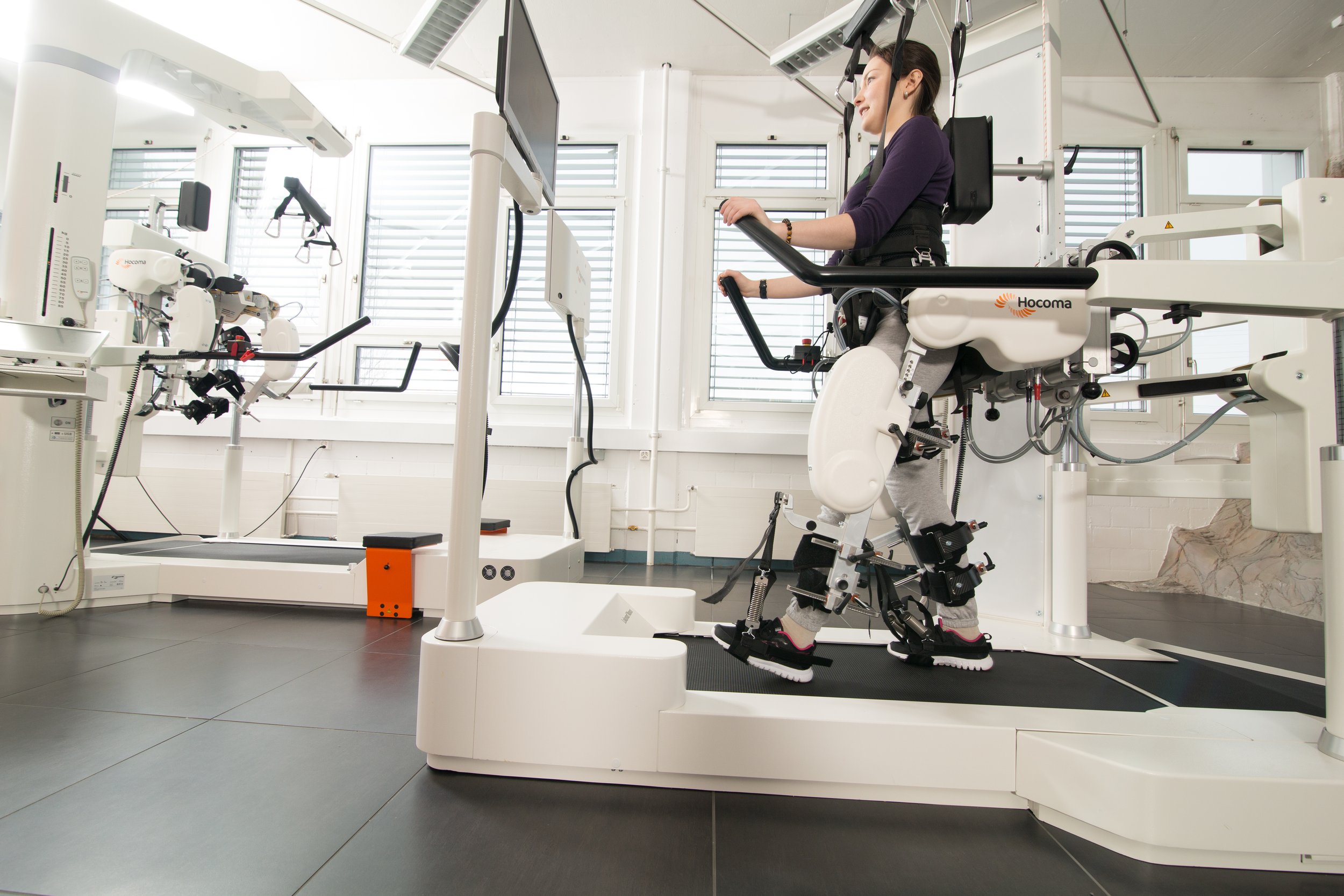 A woman with a medical or robotic device on her legs, seated on a specialized machine in a clinical setting, with monitors and equipment around.