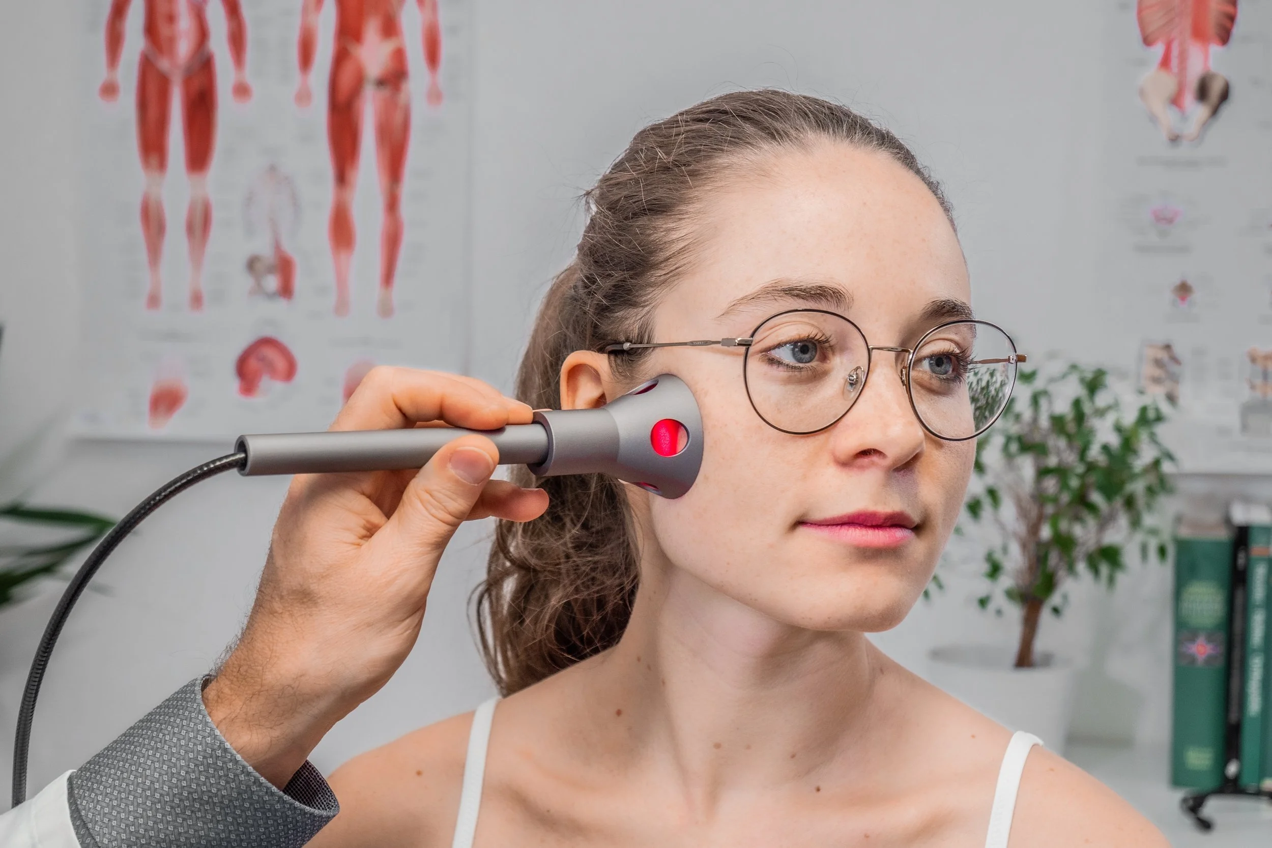 Woman with glasses receiving a medical or cosmetic treatment on her face in a clinical setting with anatomical posters in the background.