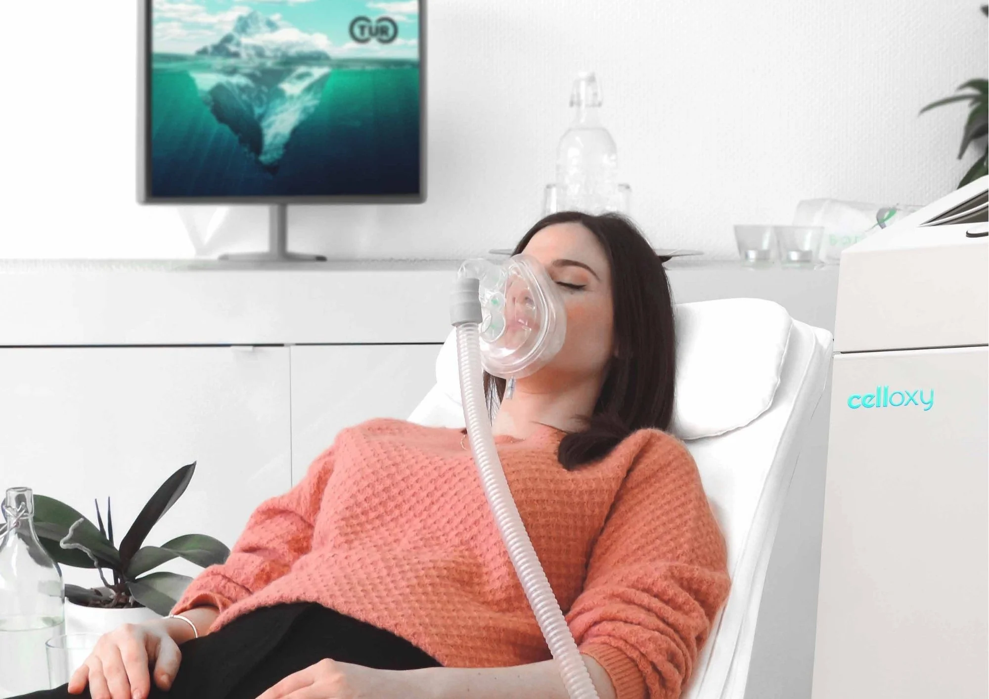 A woman with dark hair wearing an orange sweater and gas mask, lying in a white chair in a medical or clinical setting, with a monitor displaying underwater footage and a potted plant nearby.