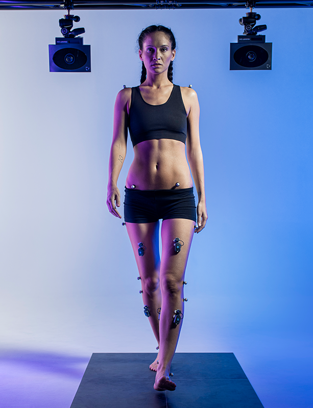 A woman in black athletic wear walking barefoot on a black platform, with her body attached to sensors for a motion study or dance analysis, against a plain background with studio lighting.