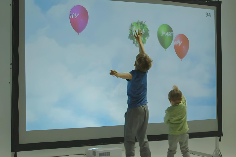 Two children are standing in front of a projector screen, reaching toward animated balloons labeled 'HAPPY' and 'PARTY' against a sky background.