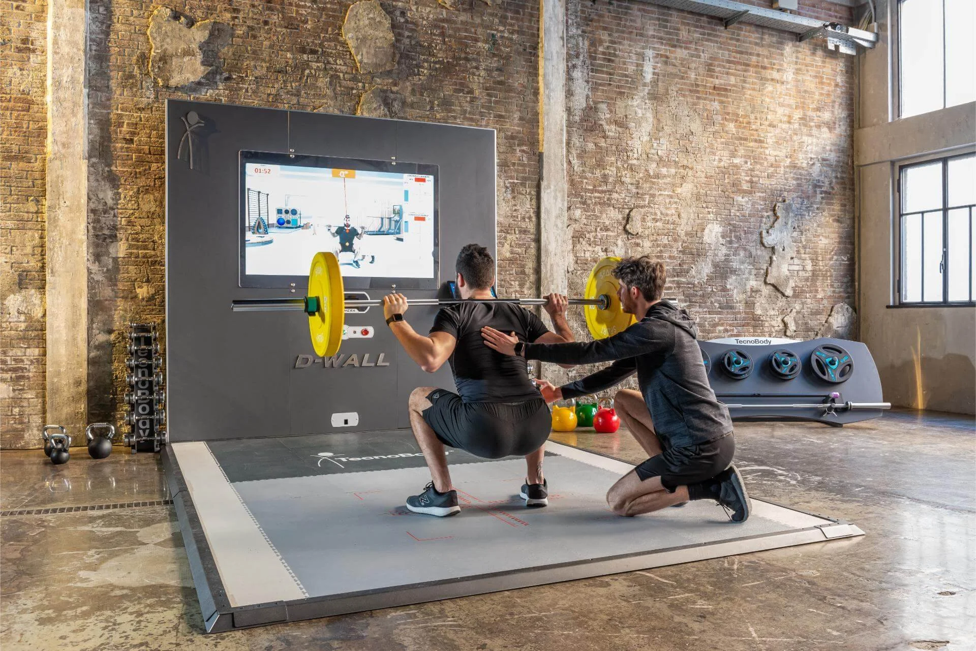 Two men are engaged in a weightlifting exercise in a gym, with one person squat-lifting a barbell while the other person is spotting him. They are on a gray workout platform in front of a large TV screen mounted on a brick wall. The gym has kettlebells, colorful medicine balls, and fitness equipment around, with a rustic brick wall and large windows in the background.