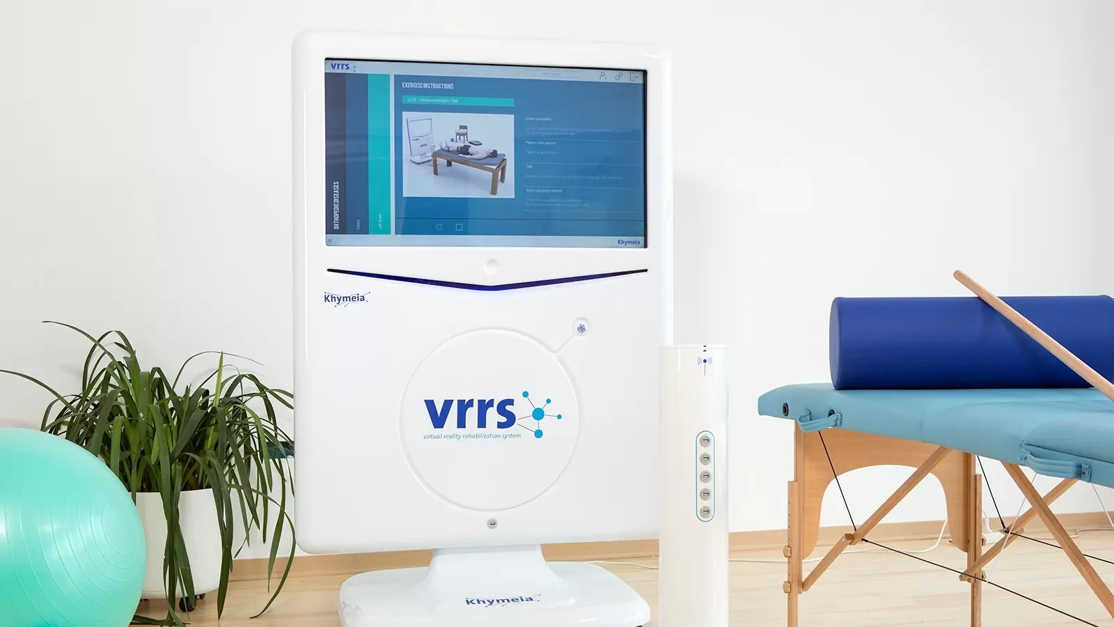 A medical rehabilitation room with a large diabetes VRT system monitor, a potted plant, a green exercise ball, and an empty padded treatment table with a roll of blue padding and a wooden stick on top.