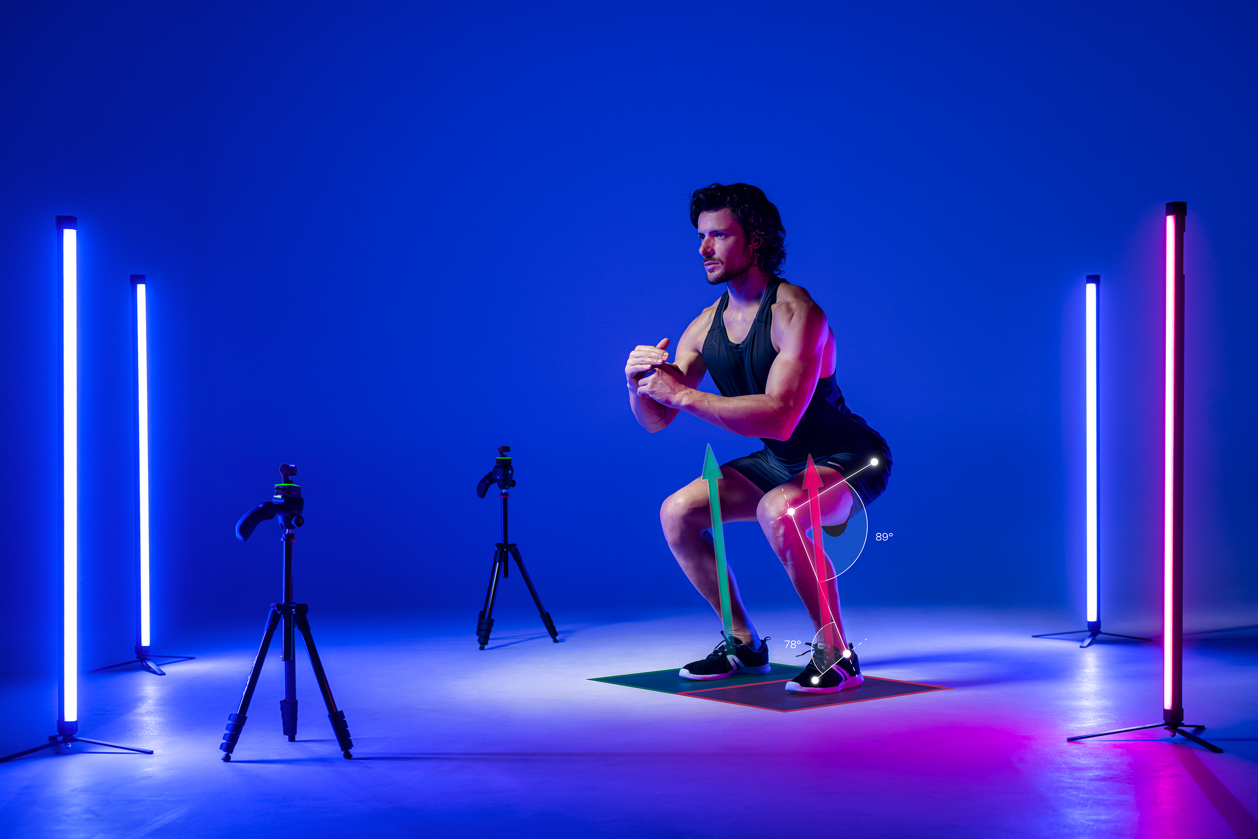 A man in athletic clothing performing a squat exercise on a mat in a room illuminated with neon lights. The room has multiple camera tripods and a digital overlay showing angle measurements of his knee and leg alignment.