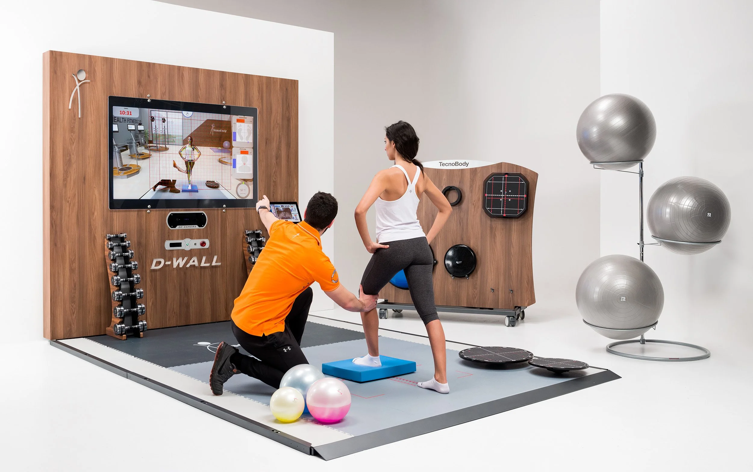 A woman performs a physical therapy move on a blue balance pad while a man, possibly a therapist, supports her. They are in a rehab or fitness setting with exercise equipment including a large screen display, dumbbells, stability balls, and wall-mounted devices. Both individuals are focused on the activity, and the environment features a white backdrop with organized fitness tools.