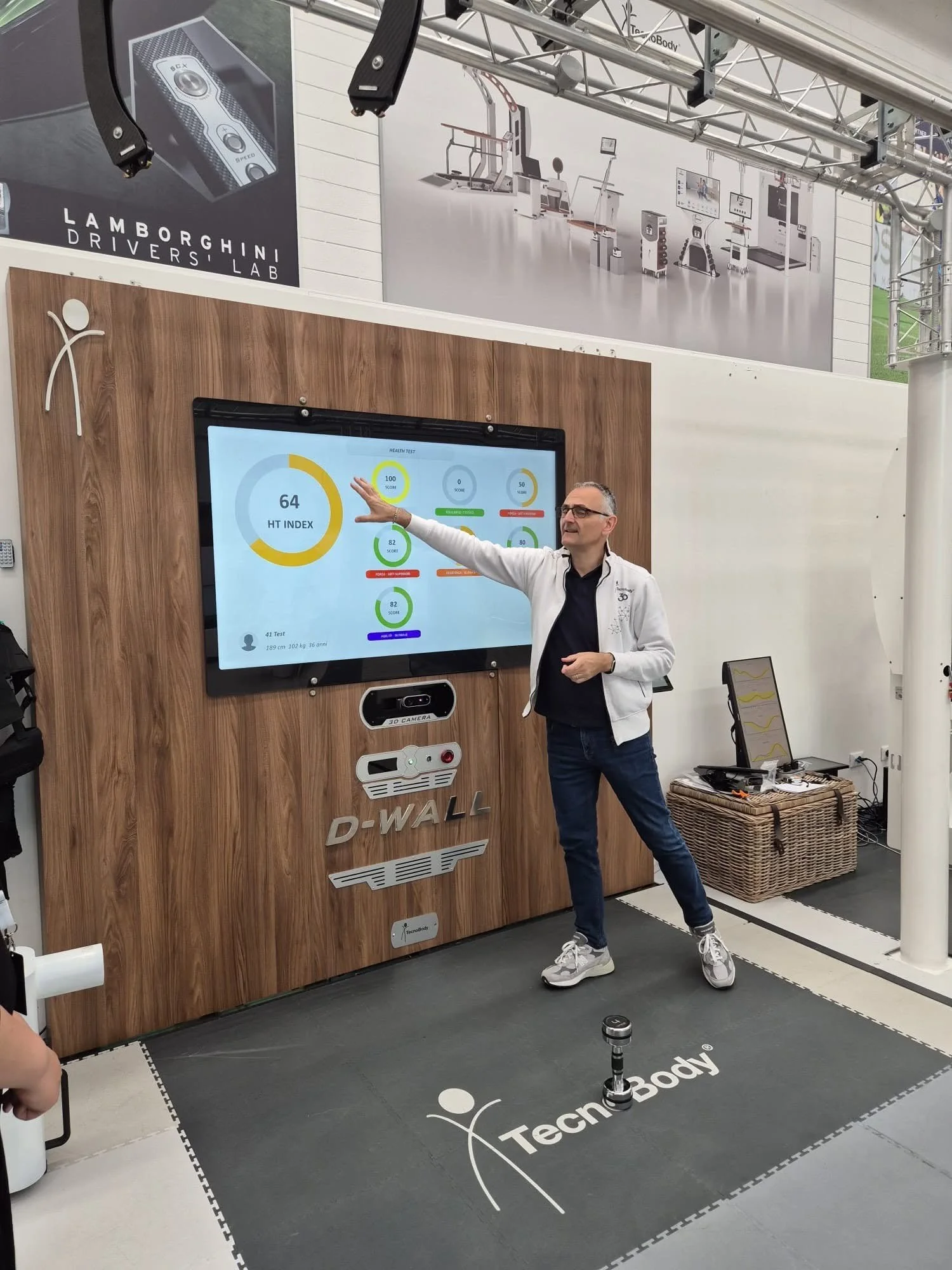 A man in glasses and a white jacket is standing in front of a large digital screen displaying health test results, in a booth with wooden paneling and TecnoBody branding. There is a black mat with the TecnoBody logo on the floor.