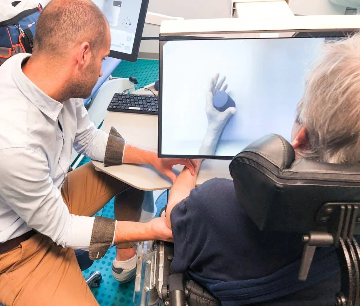 A healthcare professional is sitting at a desk holding a patient's arm, which has a bandage. A computer monitor displays an image of a hand holding a pill or capsule, with other medical equipment visible in the background.