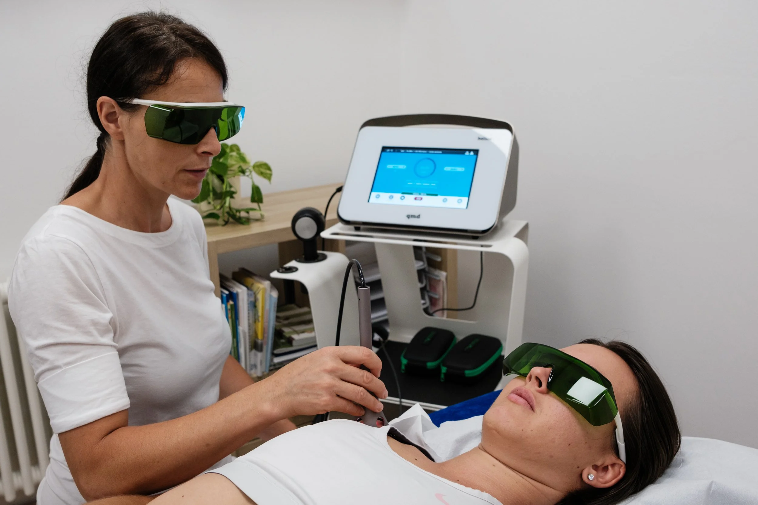 A woman wearing green eye protection goggles receives a treatment from a healthcare provider in a clinical setting, with medical equipment nearby.