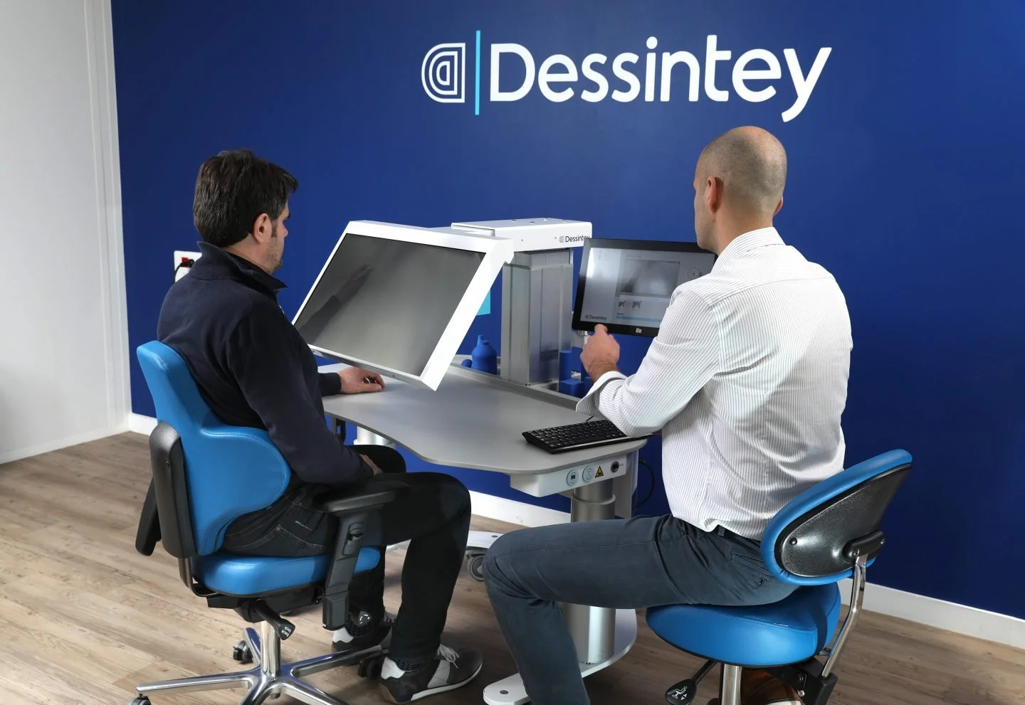 Two men sitting at a desk with computer monitors, one of them holding a tablet, in a room with a blue wall that has the logo 'Dessintey' on it.