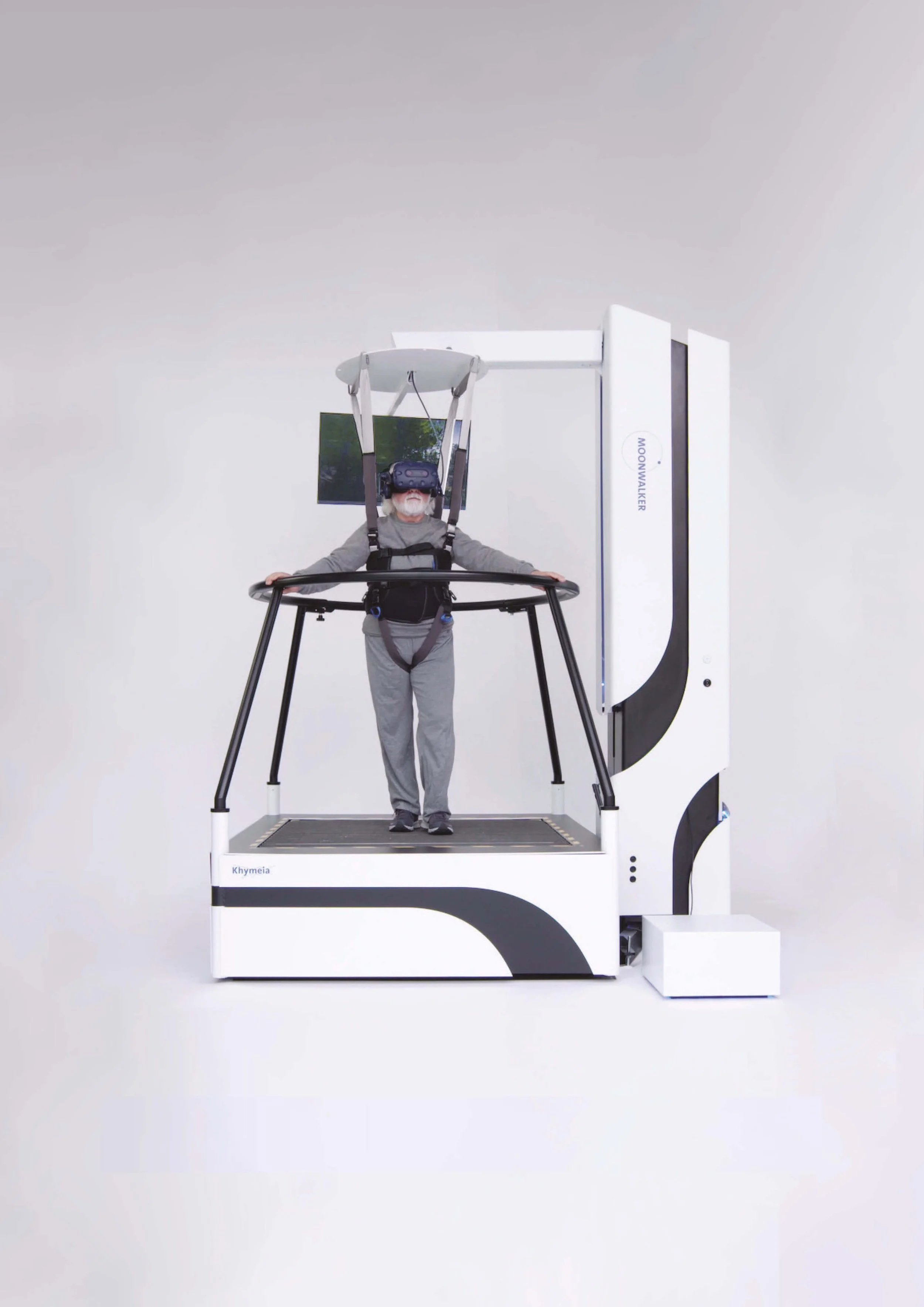 An elderly man in gray clothing stands on a platform inside a white room, wearing a virtual reality headset and harness, with a safety railing, in front of a large white machine labeled 'MOONWALKER' and 'Khymeia'.