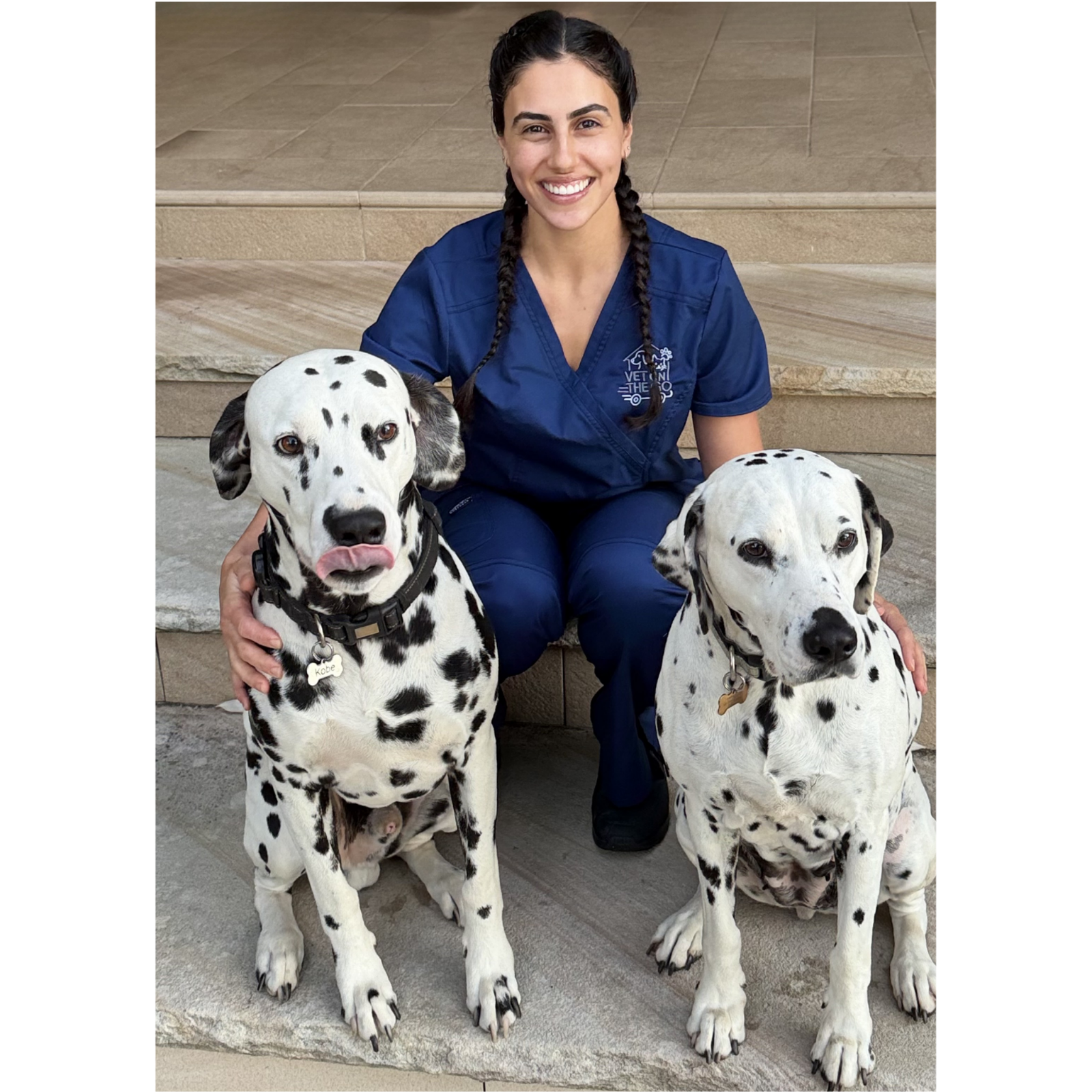 A smiling woman in veterinary scrubs sitting on steps with two Dalmatian dogs.