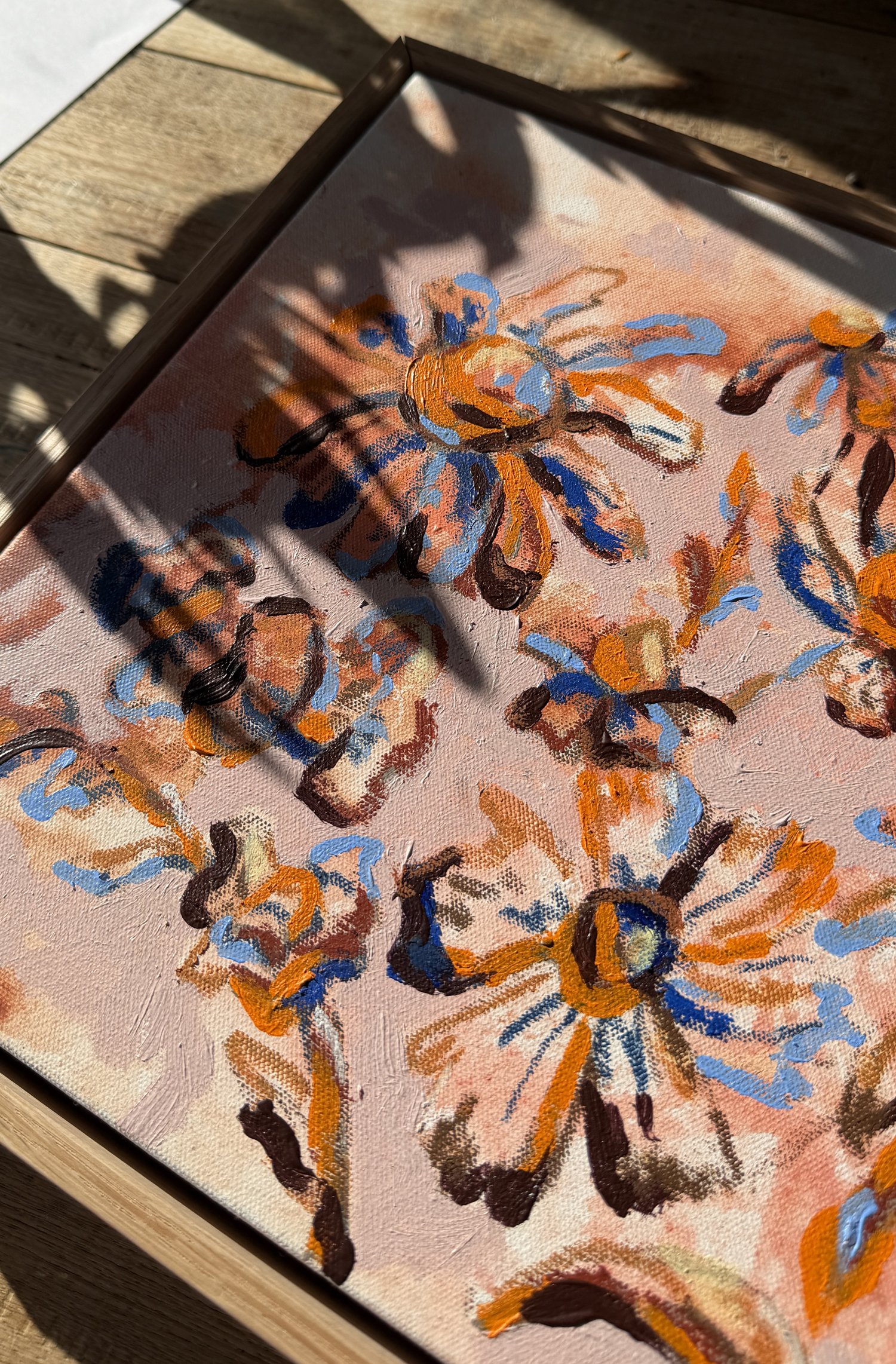 Abstract floral painting with loose orange, blue and rust-toned blooms on a blush pink background, natural oak frame