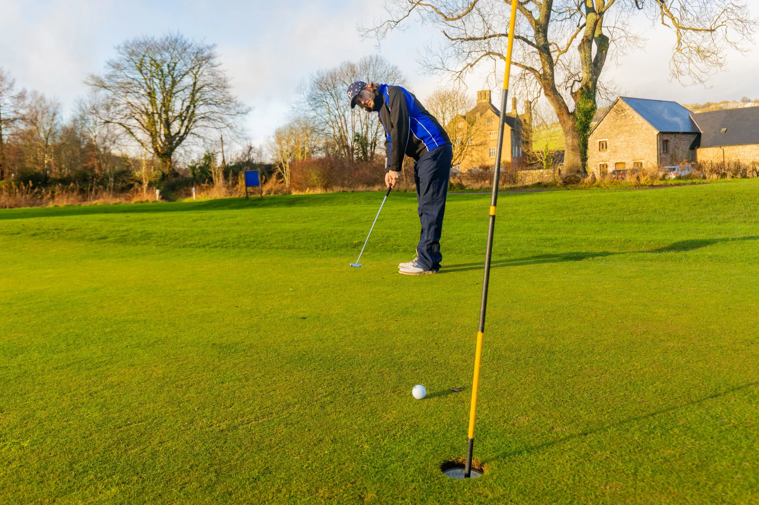 A man preparing to putt on a golf course with a flagstick nearby and a golf ball close to the hole, during a sunny day with trees and houses in the background.
