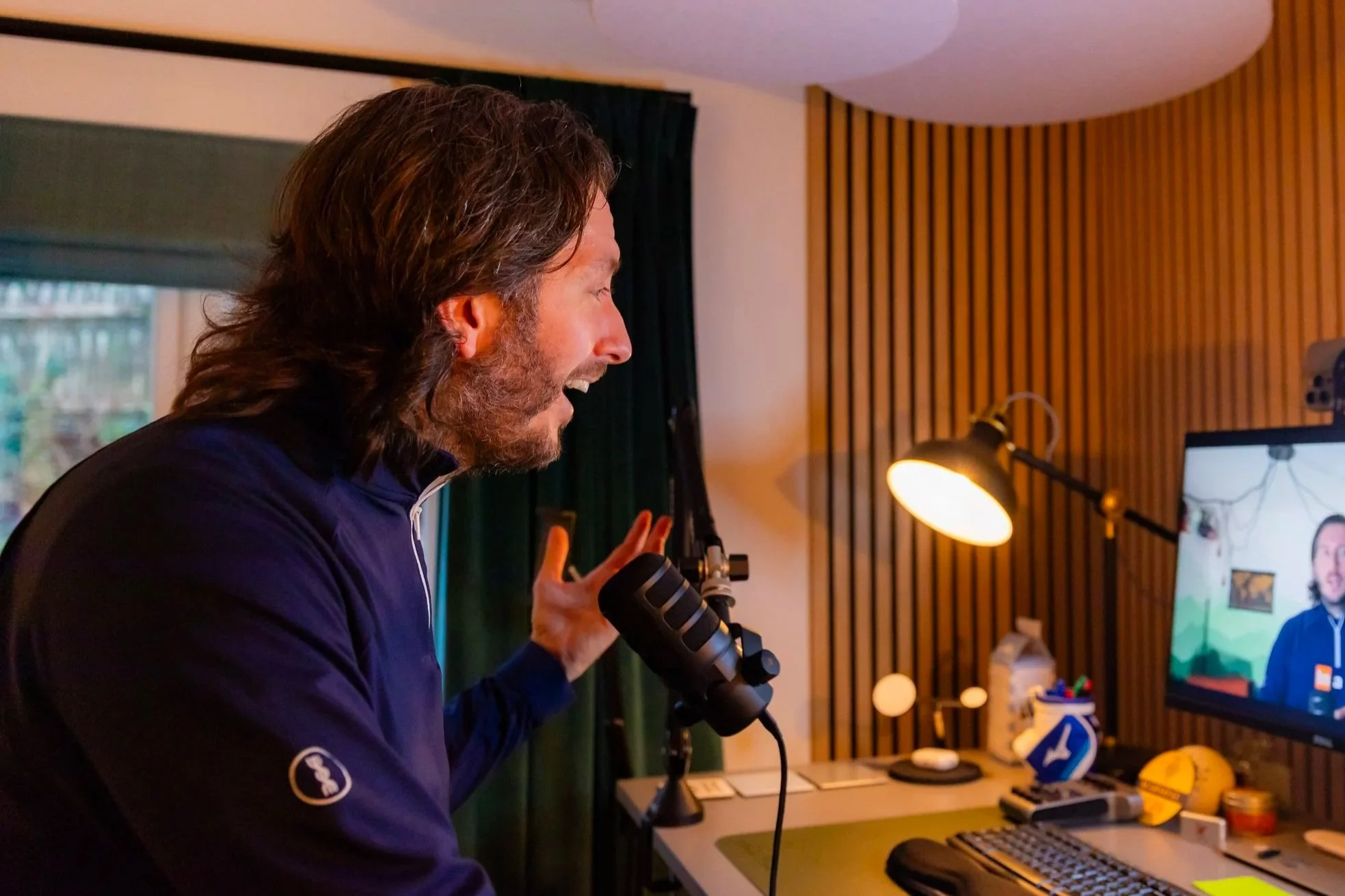 A man with long hair and a beard speaking into a microphone in front of a computer monitor, in a room with wooden wall panels and a desk lamp.