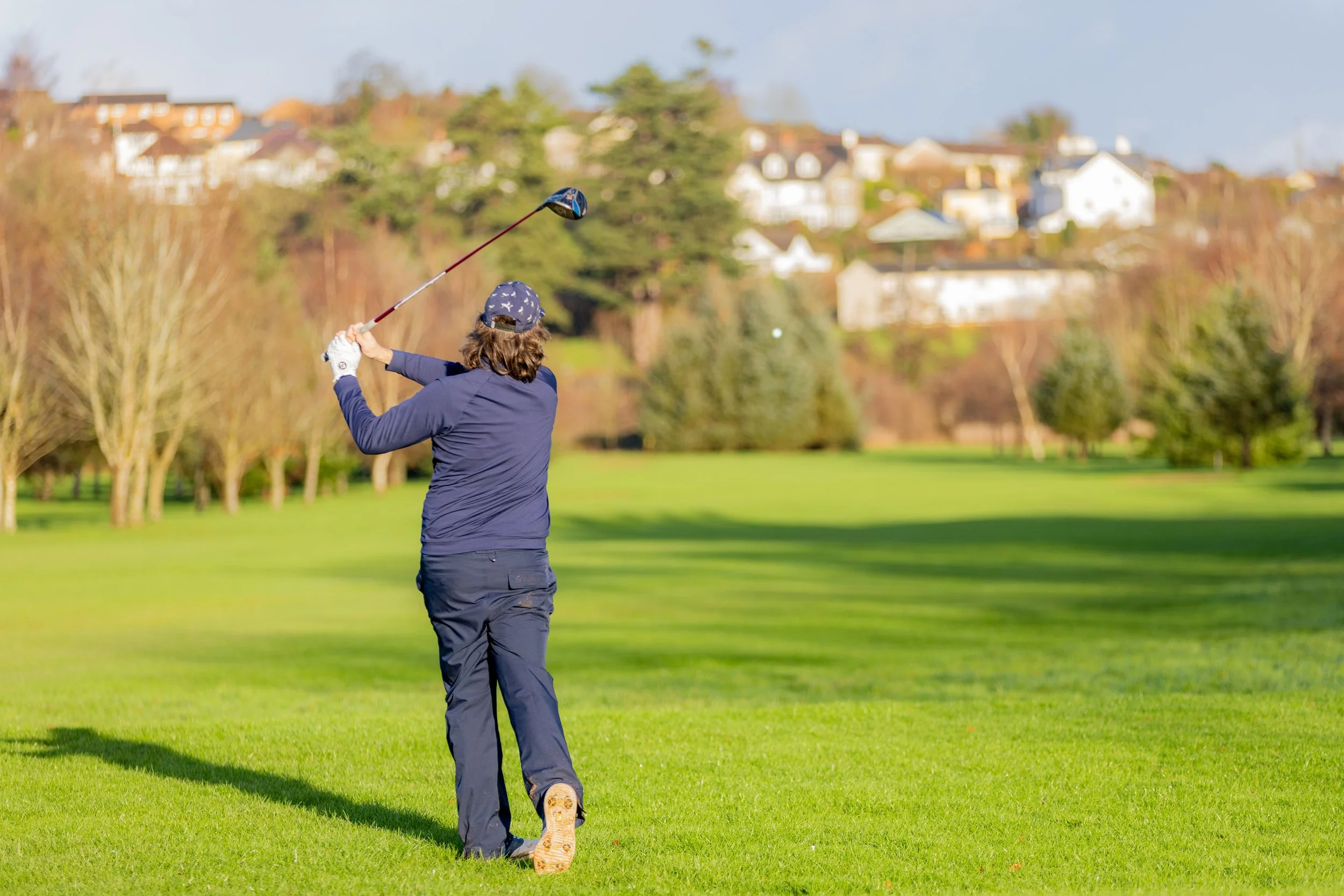 A person playing golf on a green course during daytime, swinging a golf club with an empty golf ball in the air, surrounded by trees and residential houses in the background.