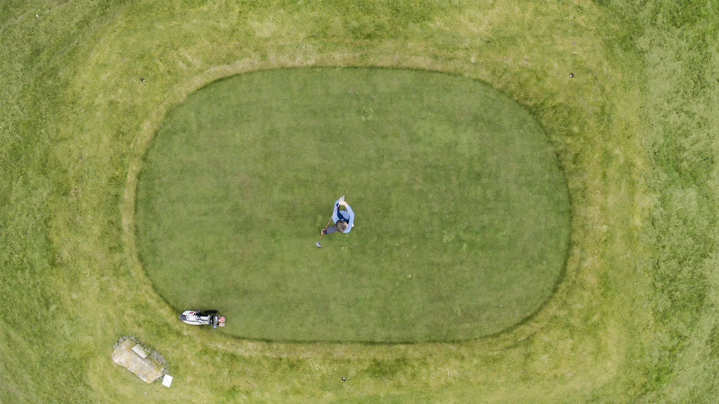 An aerial view of a small golf putting green with a golfer preparing to hit the ball, a golf bag with clubs, and a bench on the surrounding grassy area.