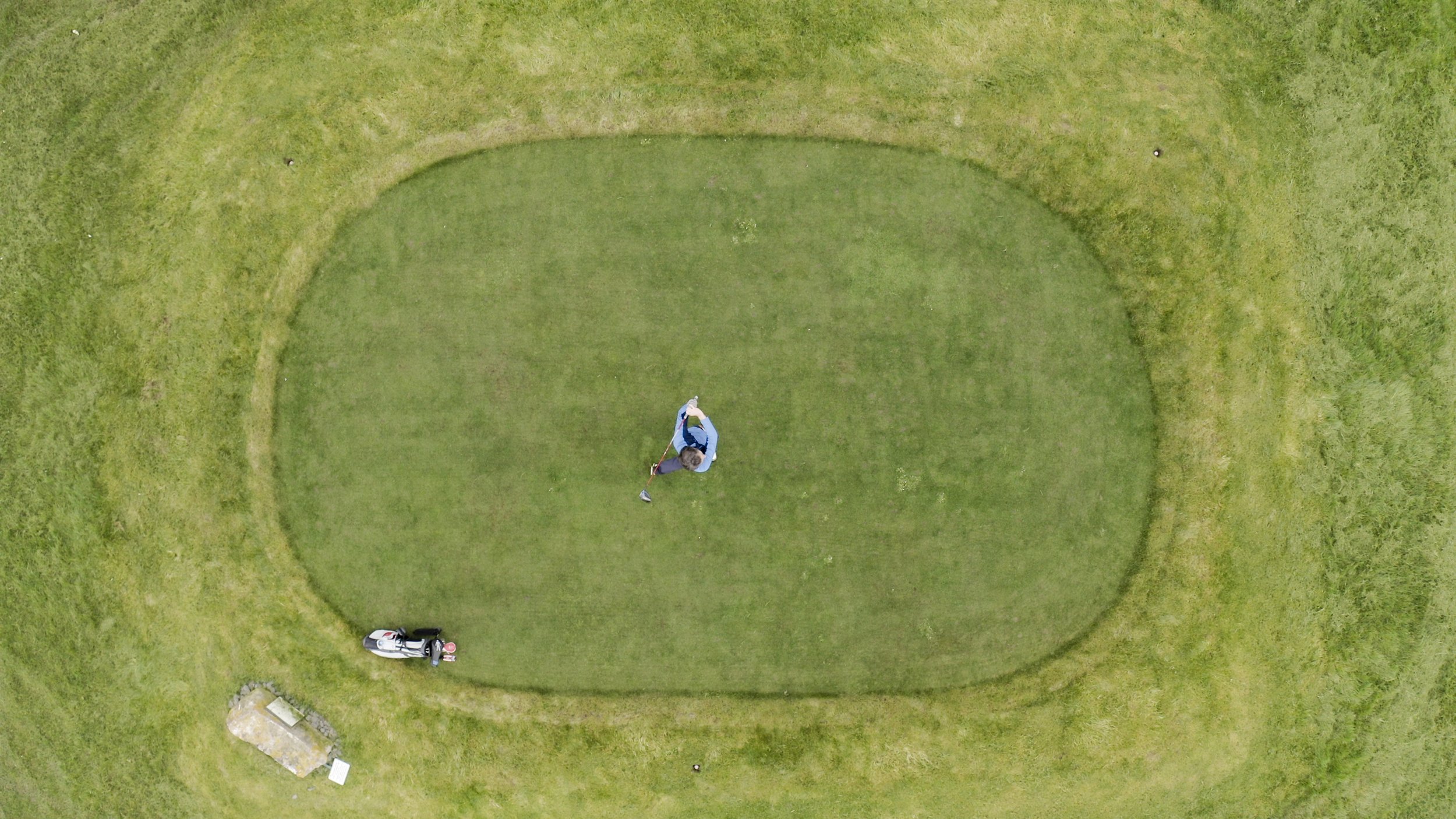 An aerial view of a man preparing to hit a golf ball on a putting green surrounded by grass, with golf clubs and a golf bag nearby.