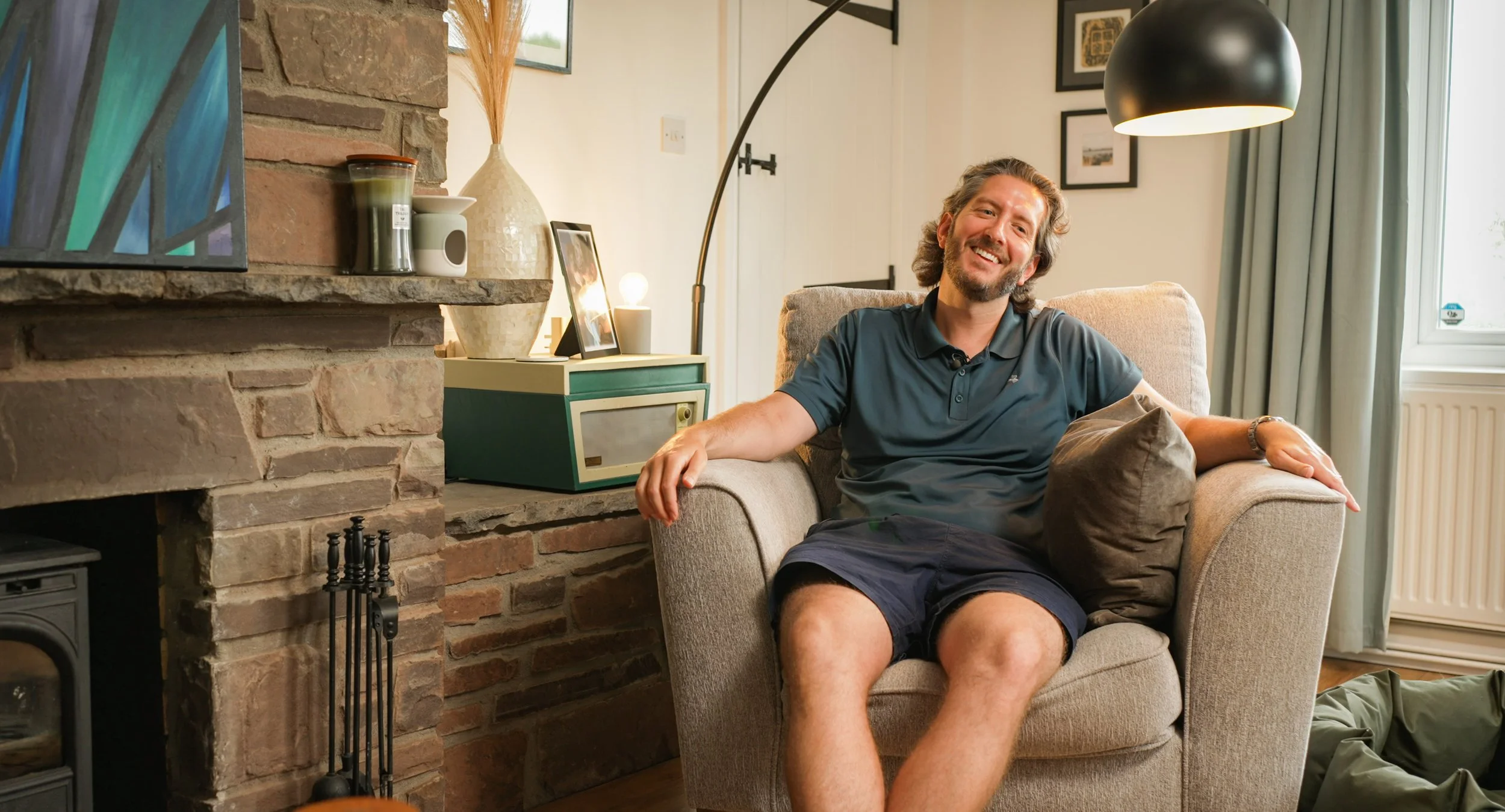 A smiling man with long hair and a beard, wearing a blue polo shirt and shorts, sits comfortably in a beige armchair in a cozy living room with a brick fireplace and a large window with curtains.