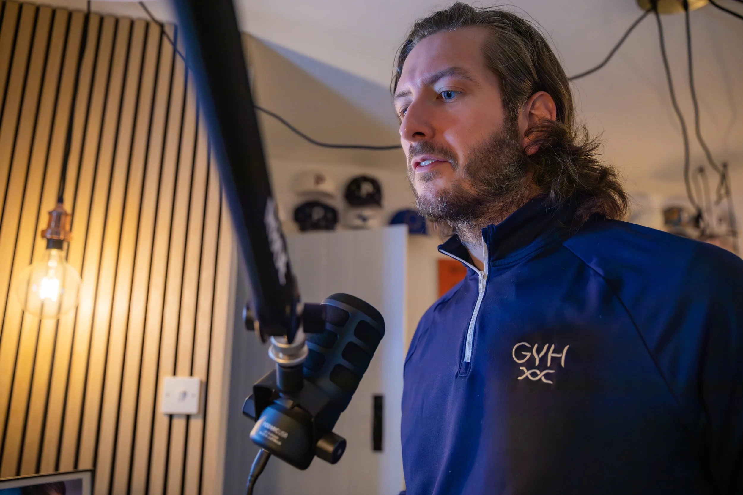 A man with long hair and a beard wearing a navy blue Gyms hoodie, speaking into a microphone in a room with wood-paneled walls and a single exposed lightbulb.