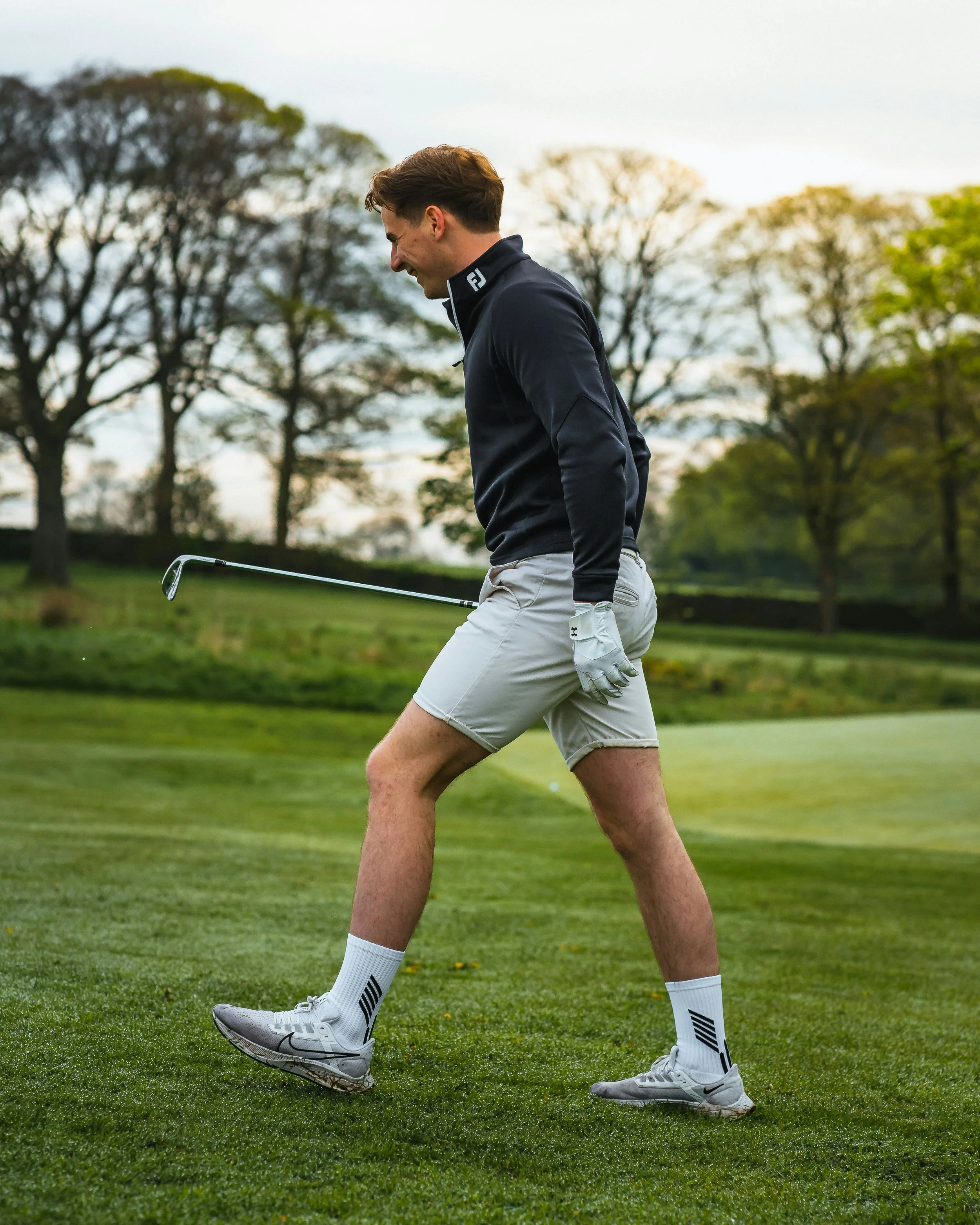 Young man on a golf course celebrating with a golf club in hand, wearing a navy jacket, light shorts, and white sports shoes.