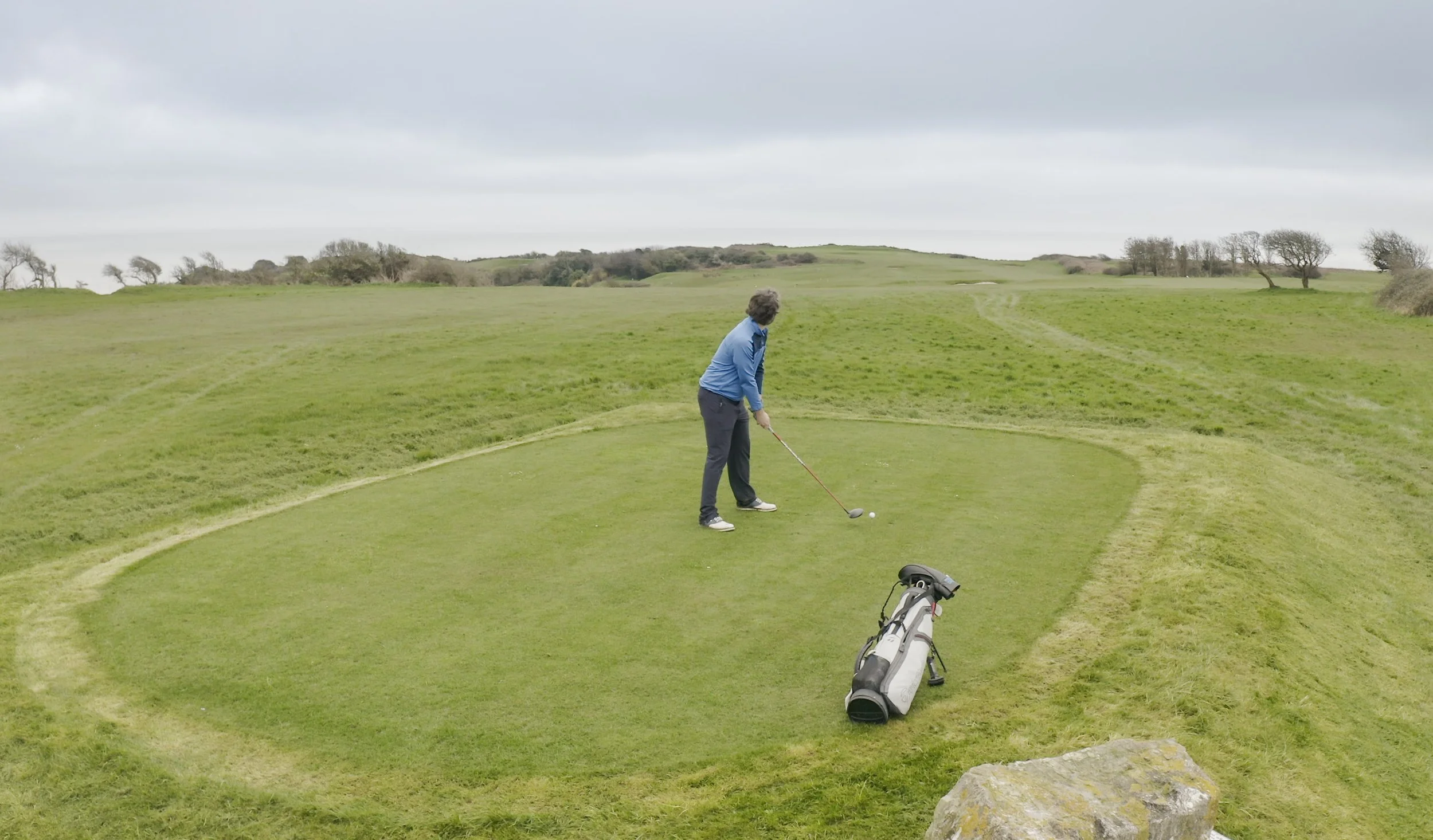 A man standing on a small golf green, preparing to hit a golf ball with a club, with a golf bag on the ground nearby, in a scenic outdoor setting with rolling grassy hills and overcast sky.