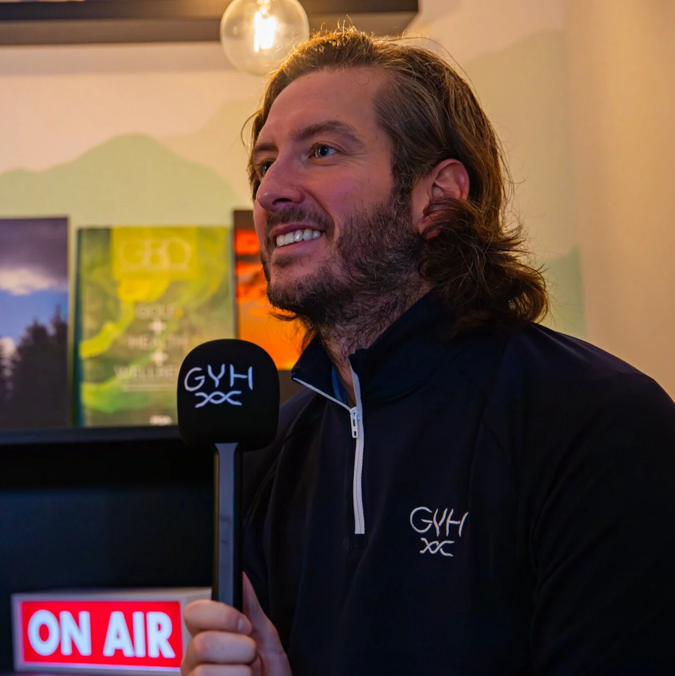 A man with long hair and beard holding a microphone labeled 'GYH' in front of a background with colorful artwork and an 'ON AIR' sign, smiling.