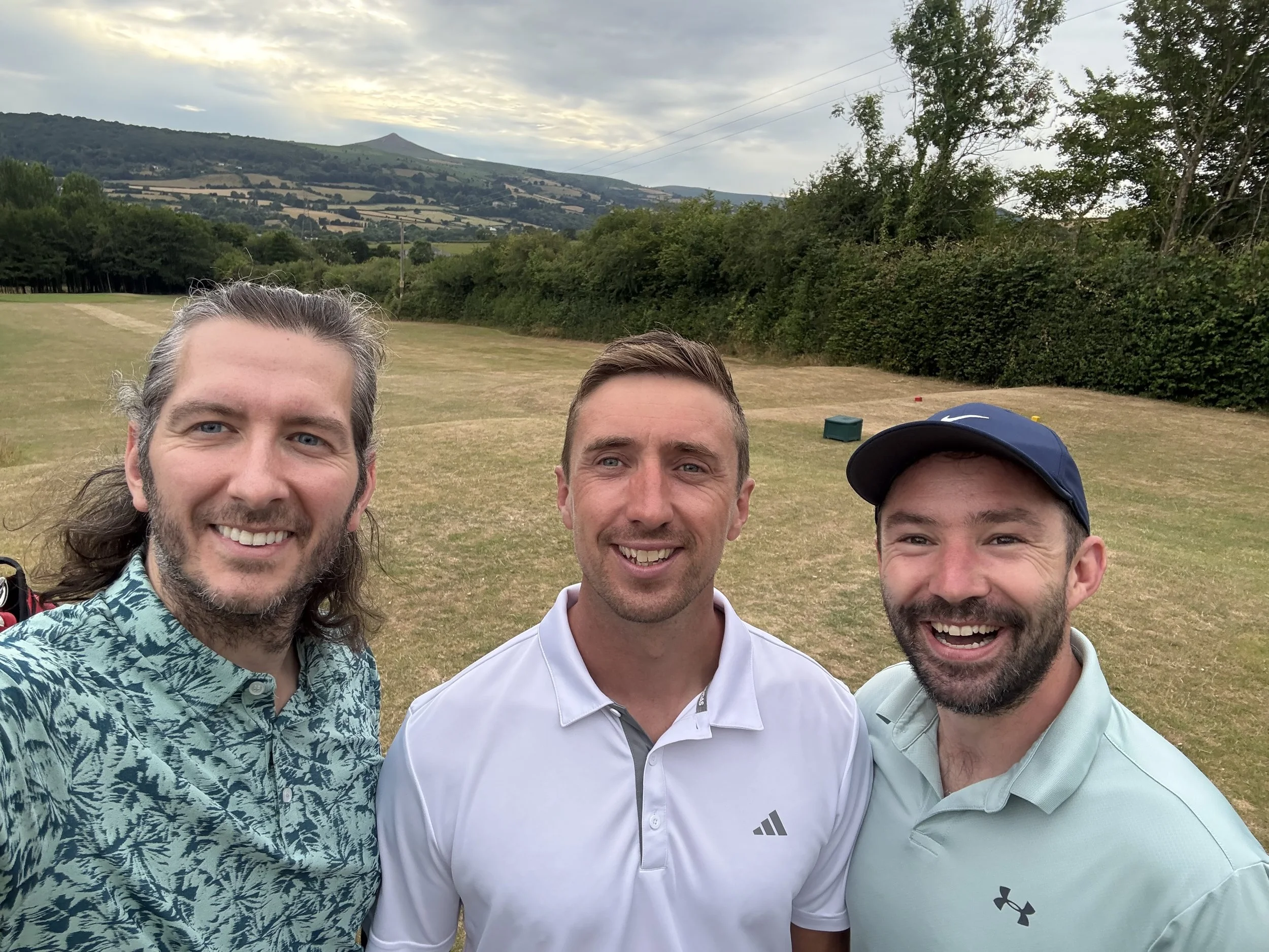 Three men smiling on a golf course with hills and trees in the background.